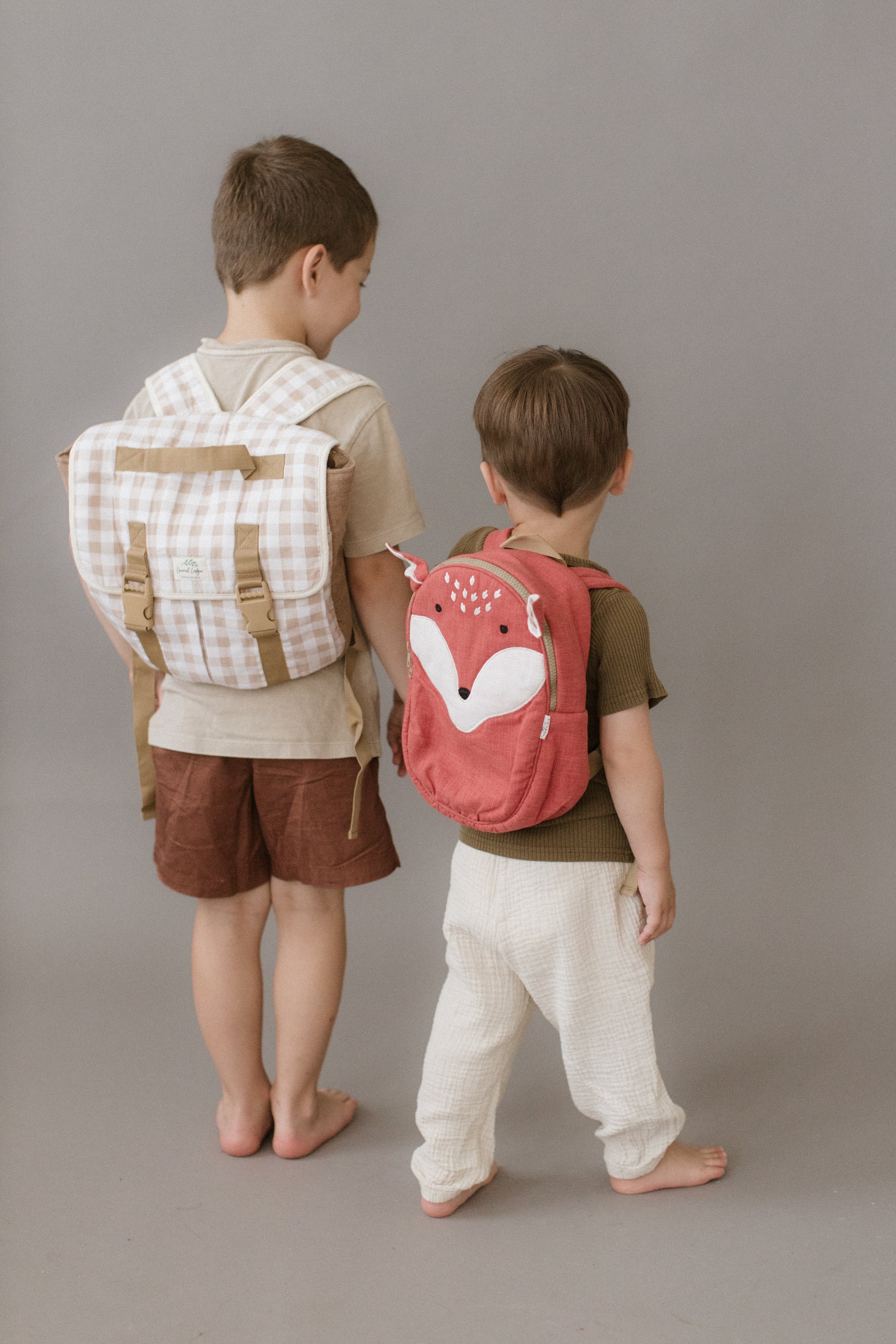 Two young boys standing barefoot against a plain gray background, facing away from the camera, each wearing backpacks. Product Photography Shoot Evanston, IL.