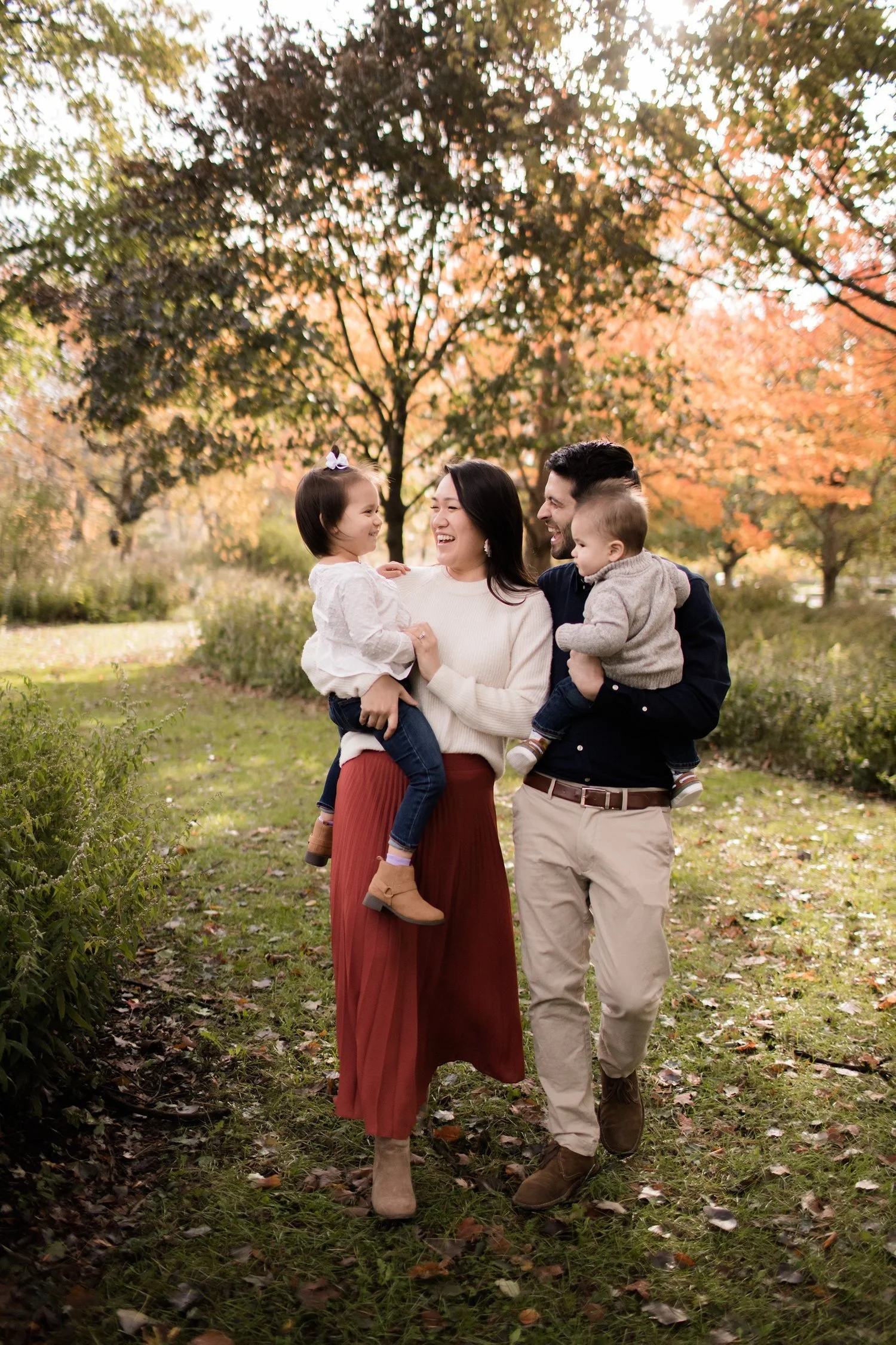 A happy family of four walking in a park during autumn, with colorful fall leaves and trees in the background. The mother and father are carrying their two young children while smiling and enjoying the moment. Family Photography Evanston IL