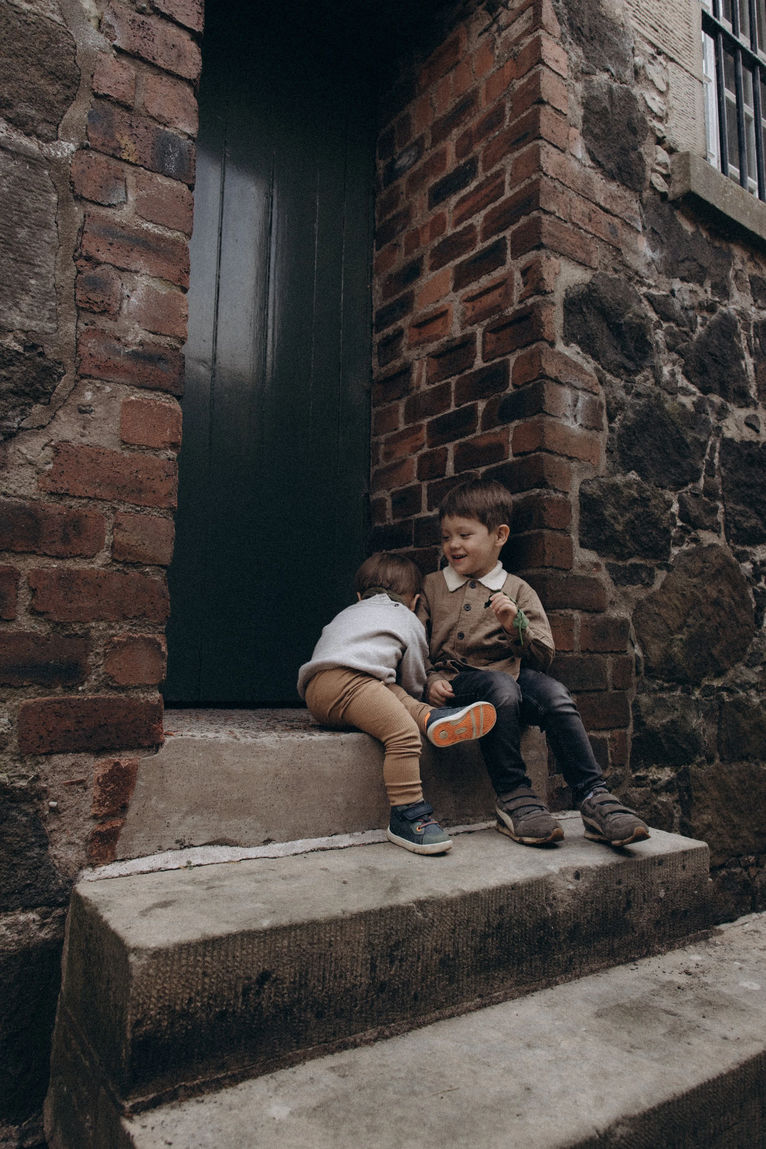 Two young children sitting on a step outside a brick and stone building, smiling and interacting with each other.