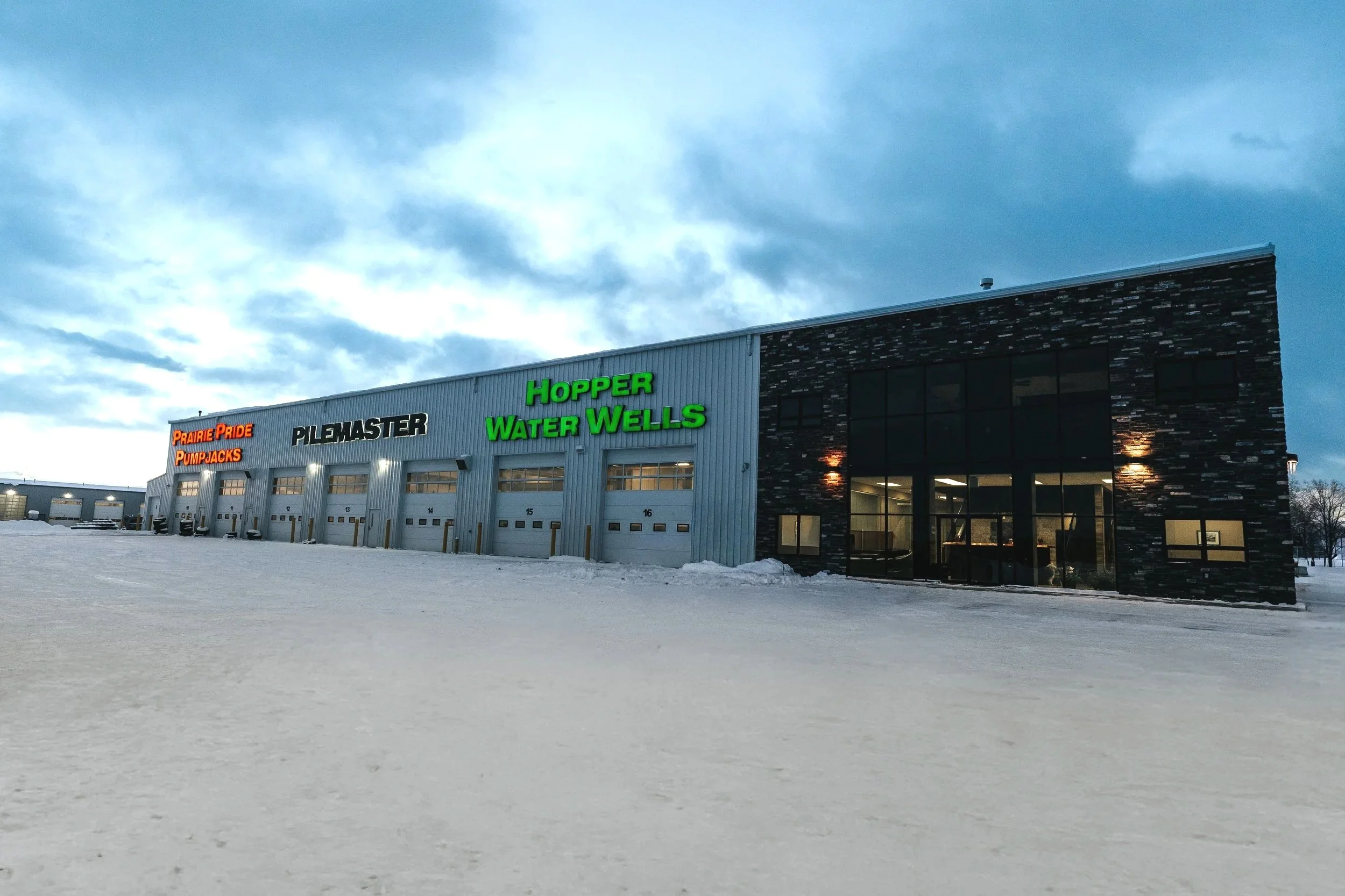 Large industrial building with colorful signs for Prairie Pride Pumpjacks, Pilemaster, Hopper Water Wells, with snowy ground and cloudy sky.