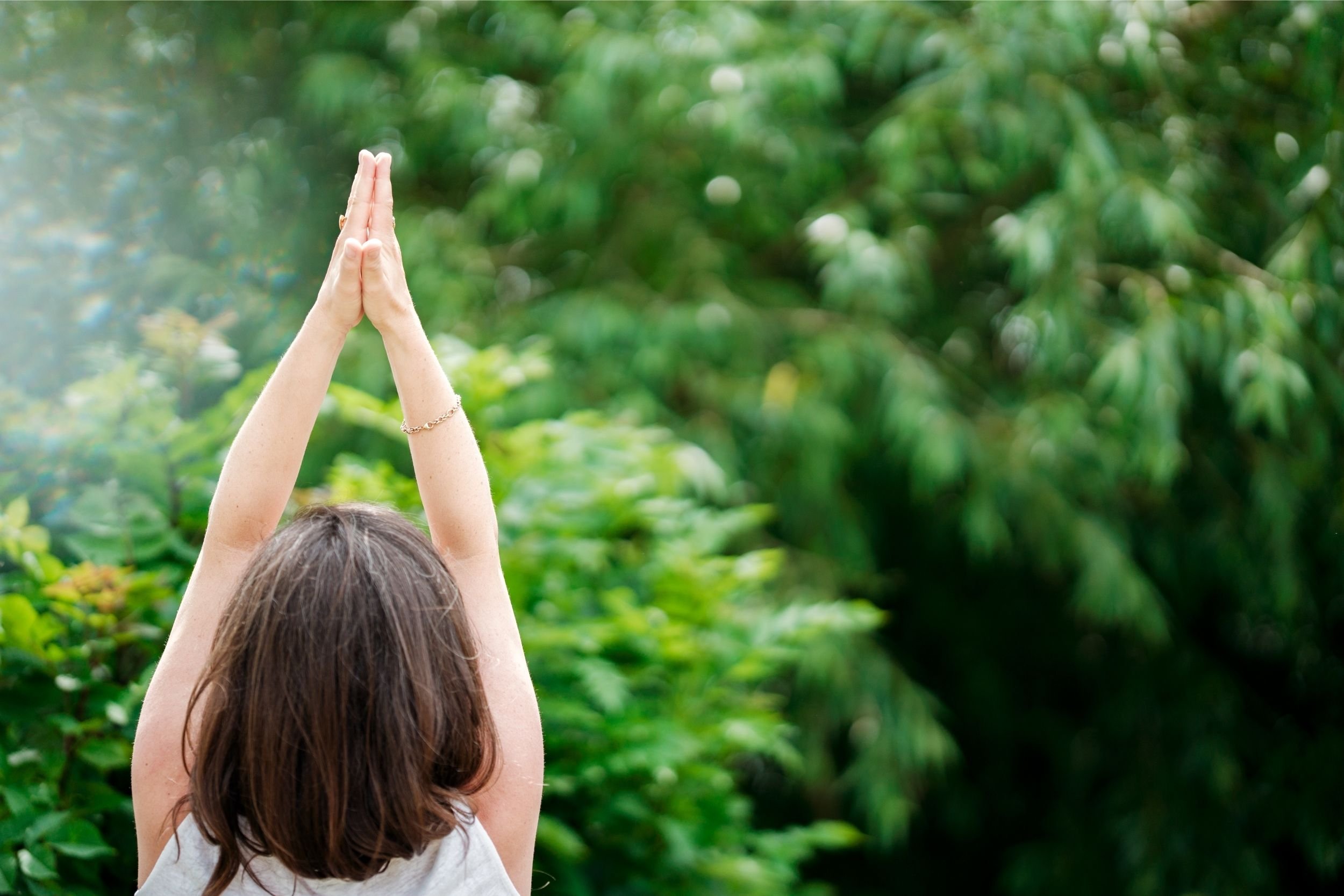 A person with long brown hair, wearing a white tank top, standing outdoors with arms raised and hands pressed together in a prayer or greeting pose, against a blurred green natural background.