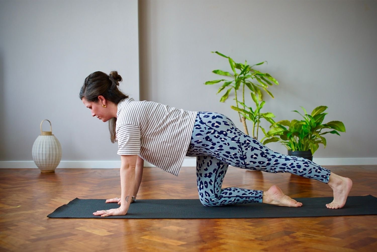 Lisa Lawton is practising yoga in a child's pose on a black mat indoors with plants and a decorative lamp in the background.