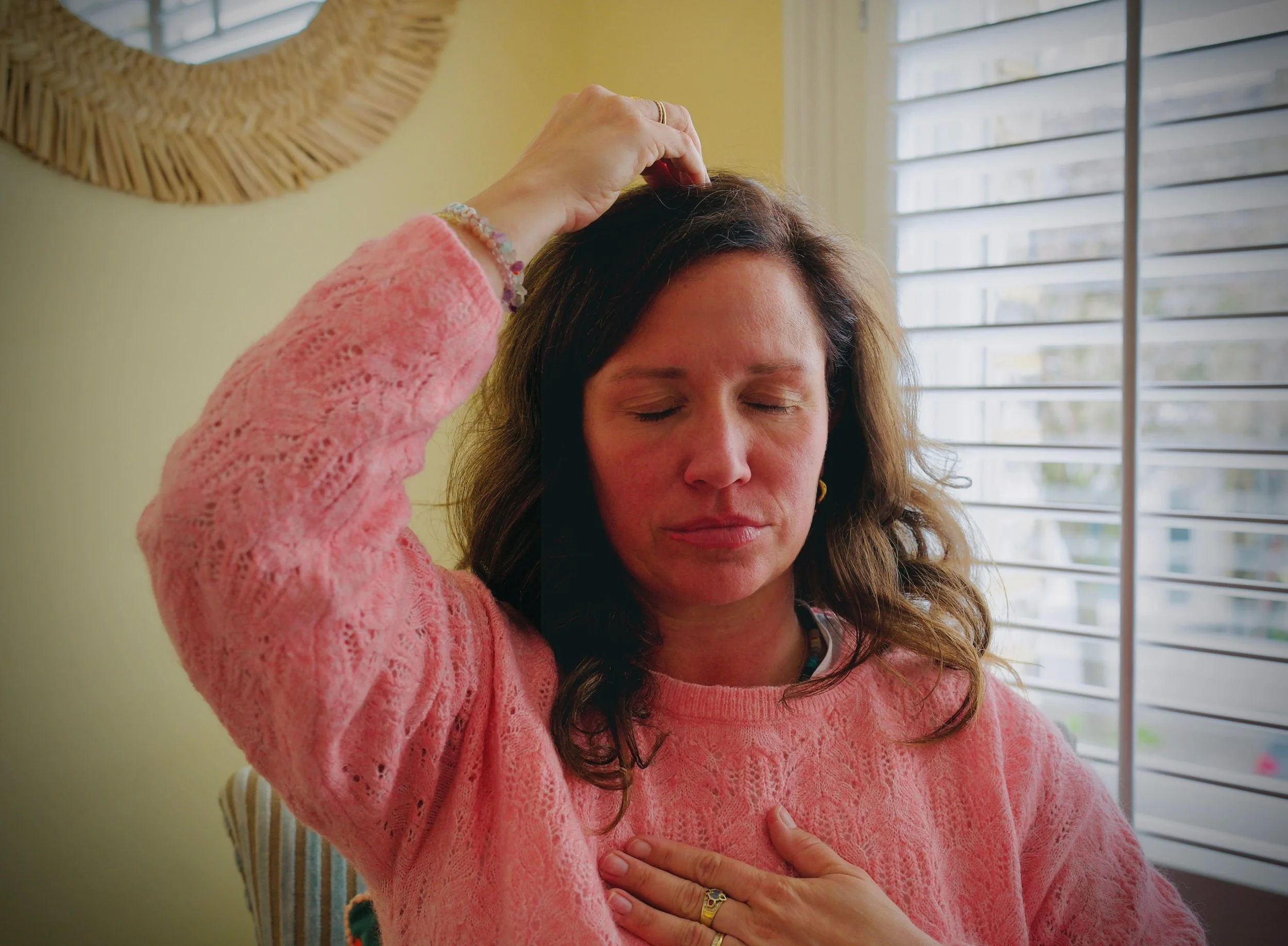 Lisa Lawton with long, wavy brown hair wearing a pink lace sweater, with her eyes closed, touching her forehead with her right hand, and resting her left hand on her chest.