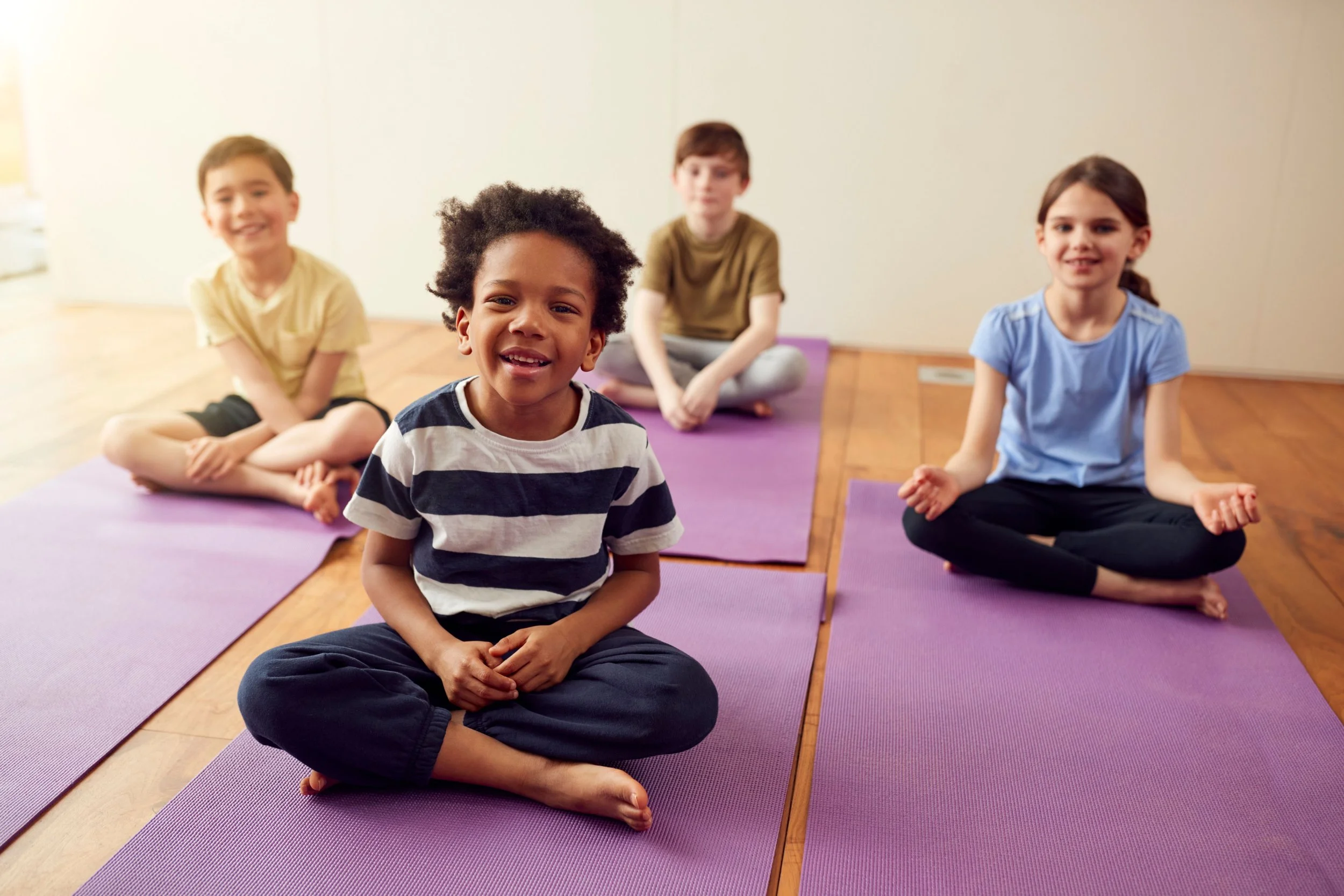 Five children sitting on purple yoga mats in a classroom, practicing yoga or meditation, with one boy smiling at the camera in the foreground.