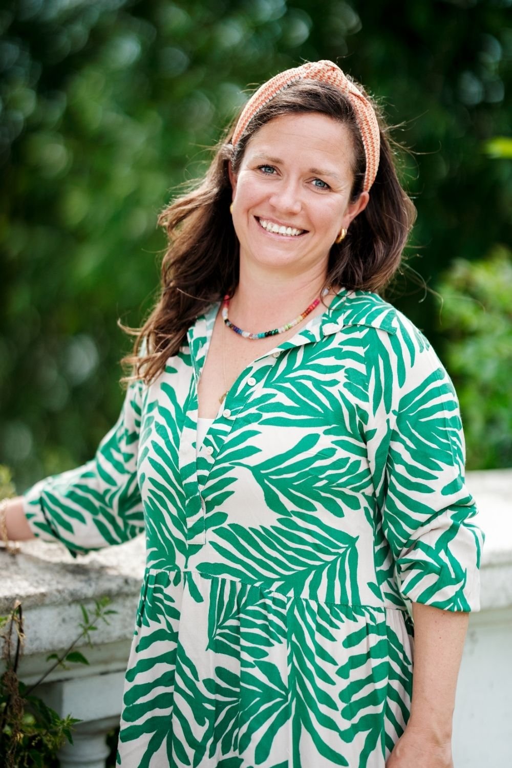 A woman with long brown hair, wearing a pink and white headband, a multicolored necklace, and a green and white leaf-patterned dress, smiles outdoors with a leafy background.