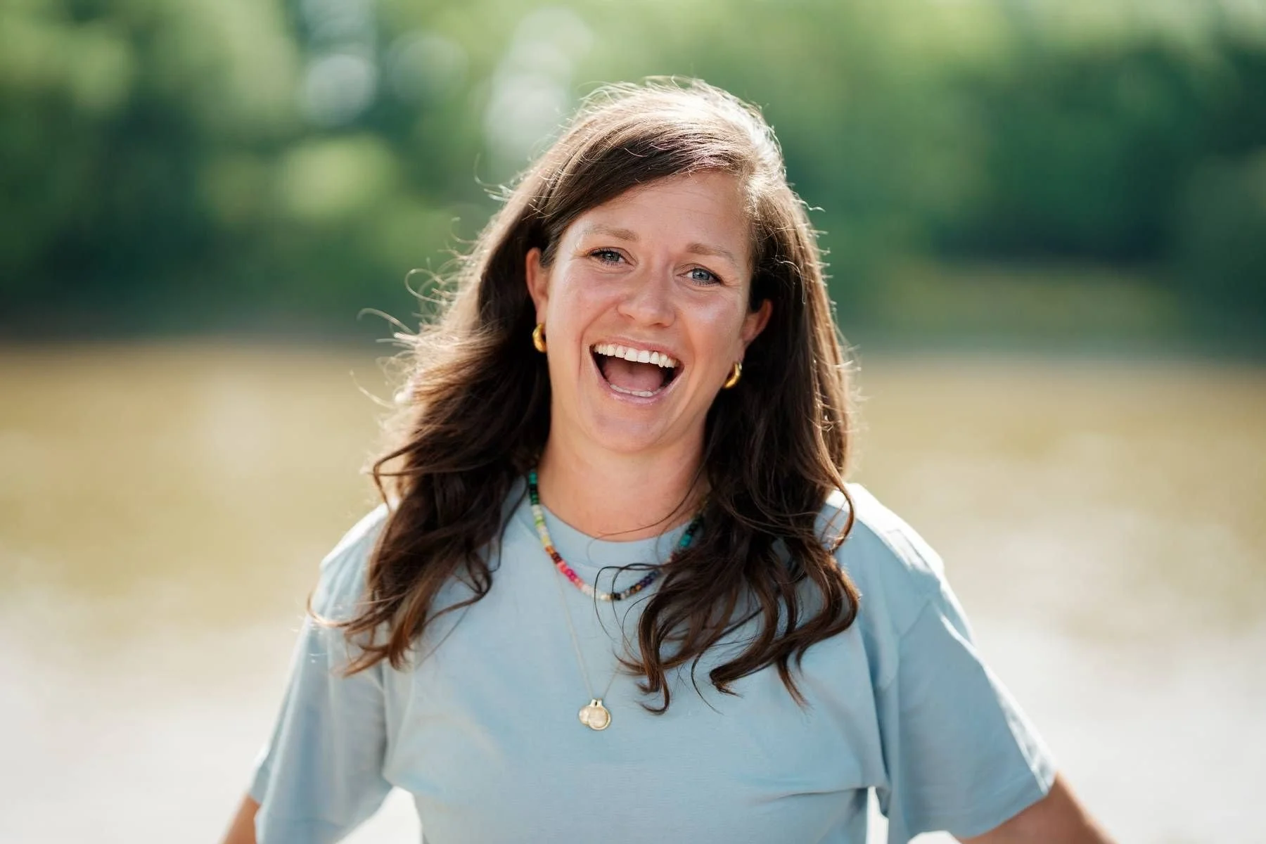 Lisa Lawton with brown hair and blue eyes smiling outdoors near a body of water, wearing a light blue shirt, colourful beaded necklaces, gold earrings, and a gold pendant necklace.