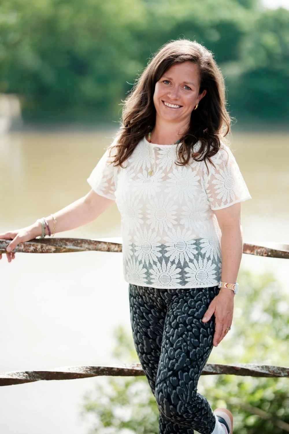 A smiling woman with brown hair, wearing a white lace flower-patterned blouse and patterned black pants, standing outdoors by a river with trees in the background.