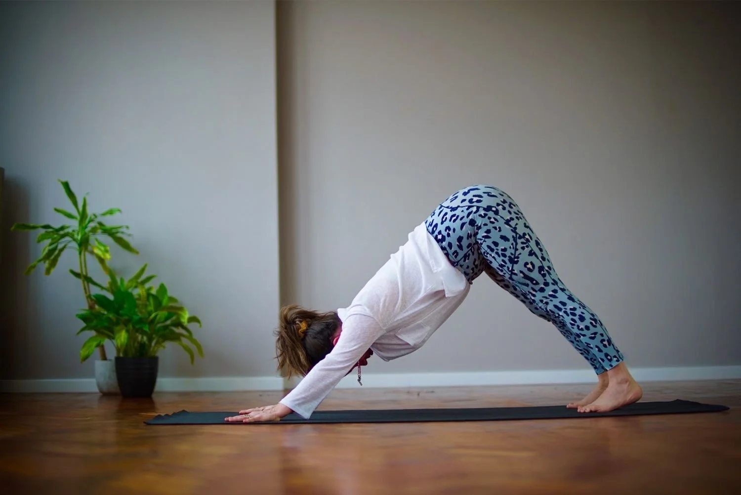 Lisa Lawton practising downward dog yoga pose on a black mat in a room with wooden flooring and bare walls, near a potted plant.