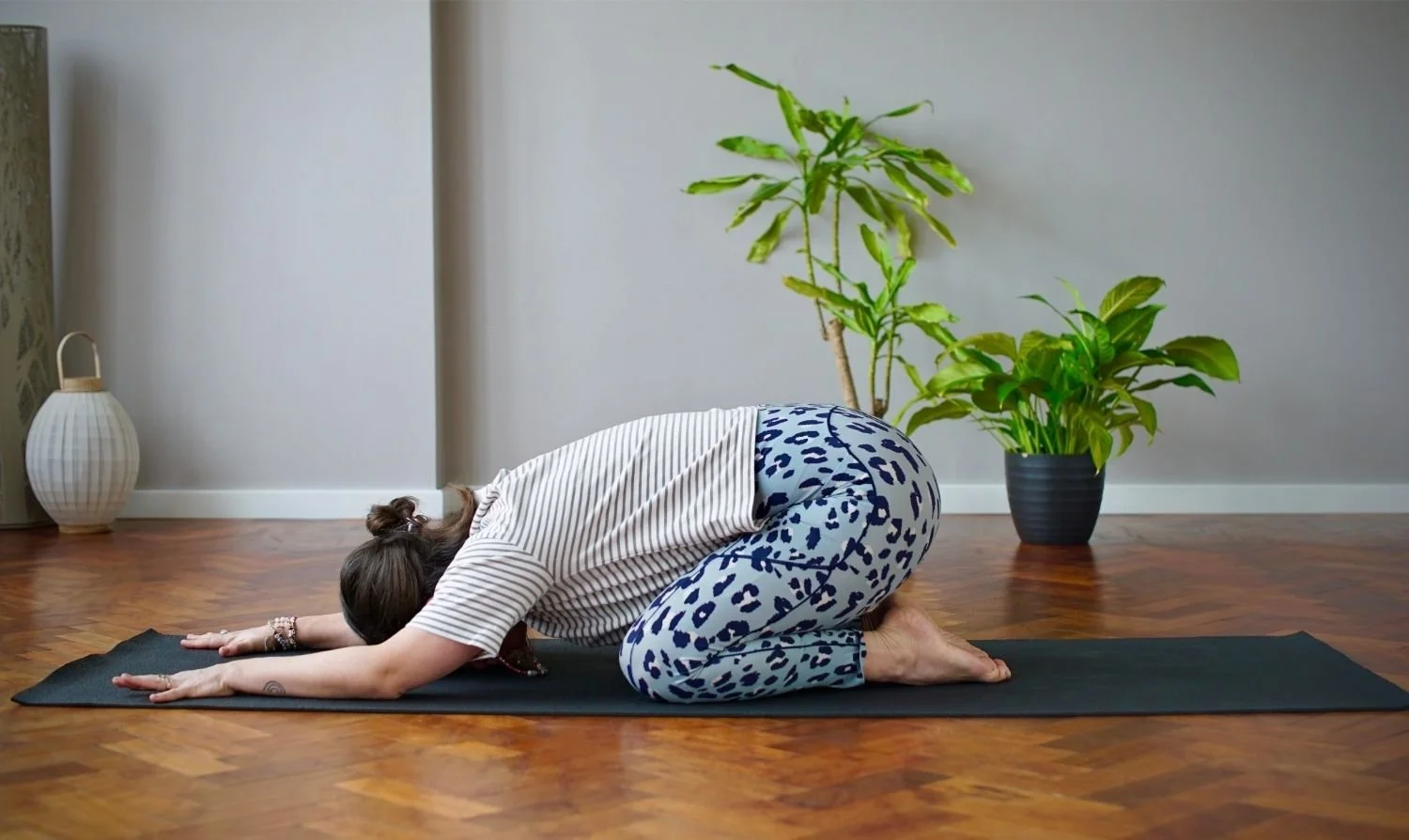 Lisa Lawton is practising yoga in child's pose on a black yoga mat in a room with wooden floors, large potted plants, and minimal decor.