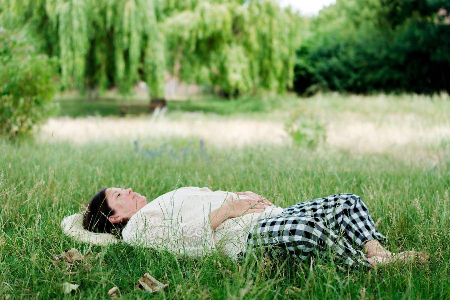 Woman lying on grass in a park near a lake, with lush green trees in the background.