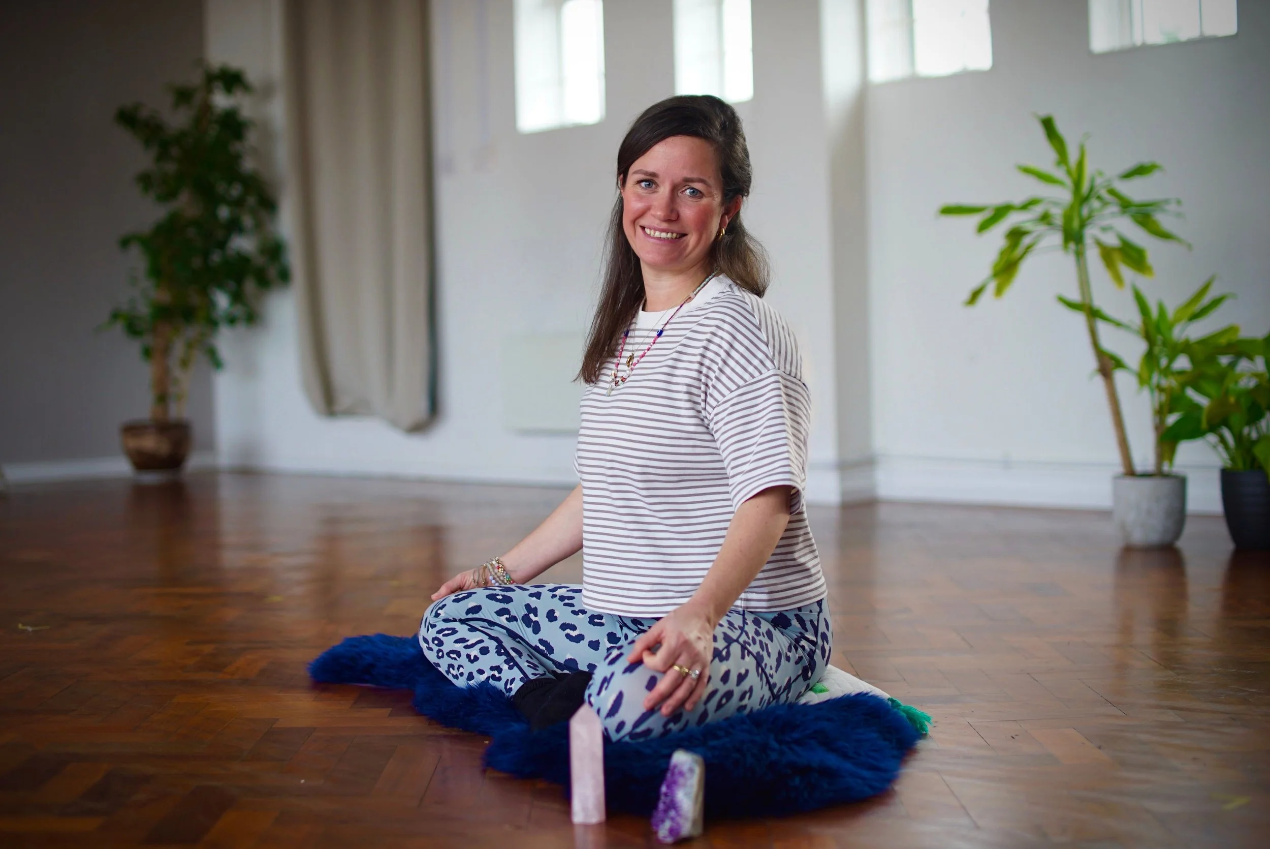 Lisa Lawton with brown hair, wearing a striped shirt and animal print pants, sitting cross-legged on a blue furry rug in a room with wooden floors and large windows, smiling at the camera.