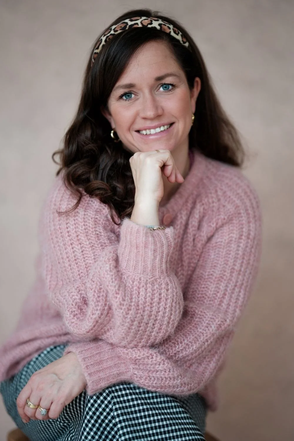 A woman with blue eyes and dark brown, curly hair wearing a pink knit sweater, leopard print headband, gold earrings, and a ring, smiling and resting her chin on her hand against a neutral background.