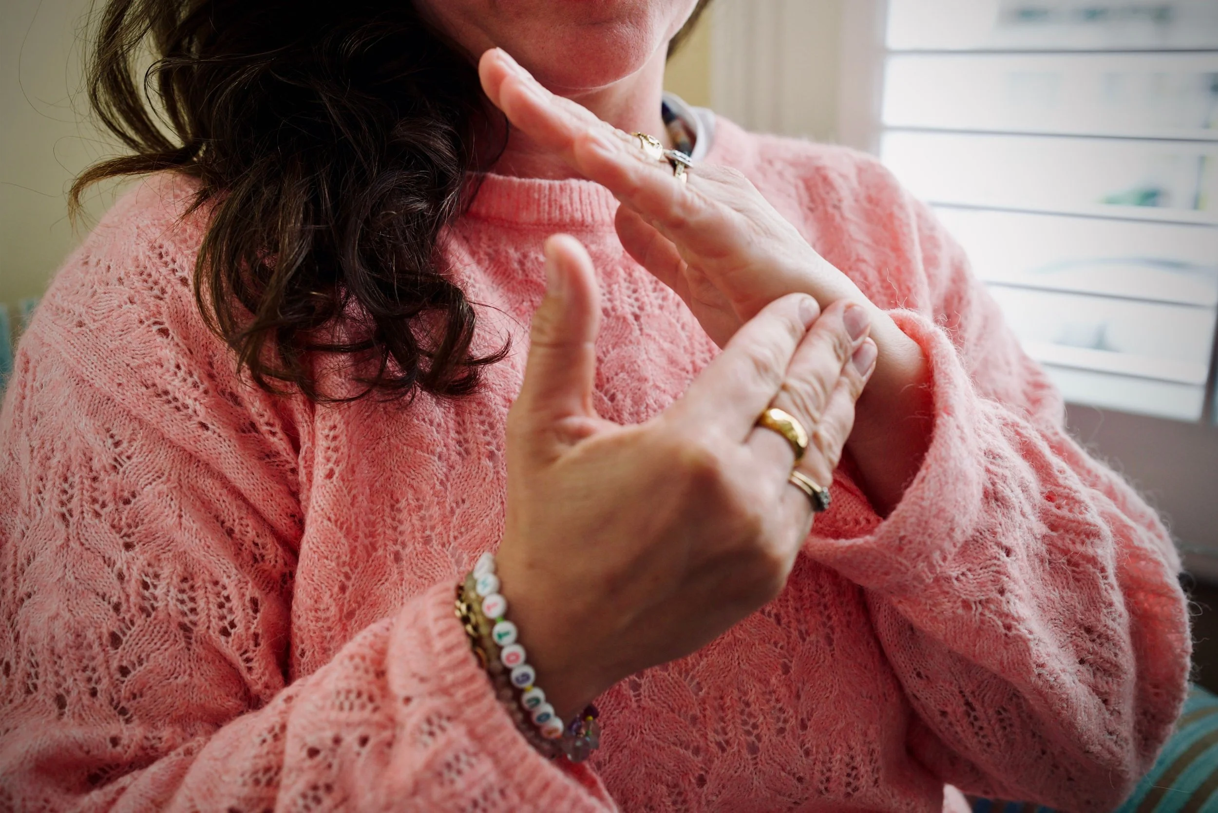 Close-up of Lisa Lawton wearing a pink, knitted sweater, touching her neck and chin with her left hand, which has rings and a colourful beaded bracelet. Her dark, curly hair partially covers her face, and she is near a window with blinds.