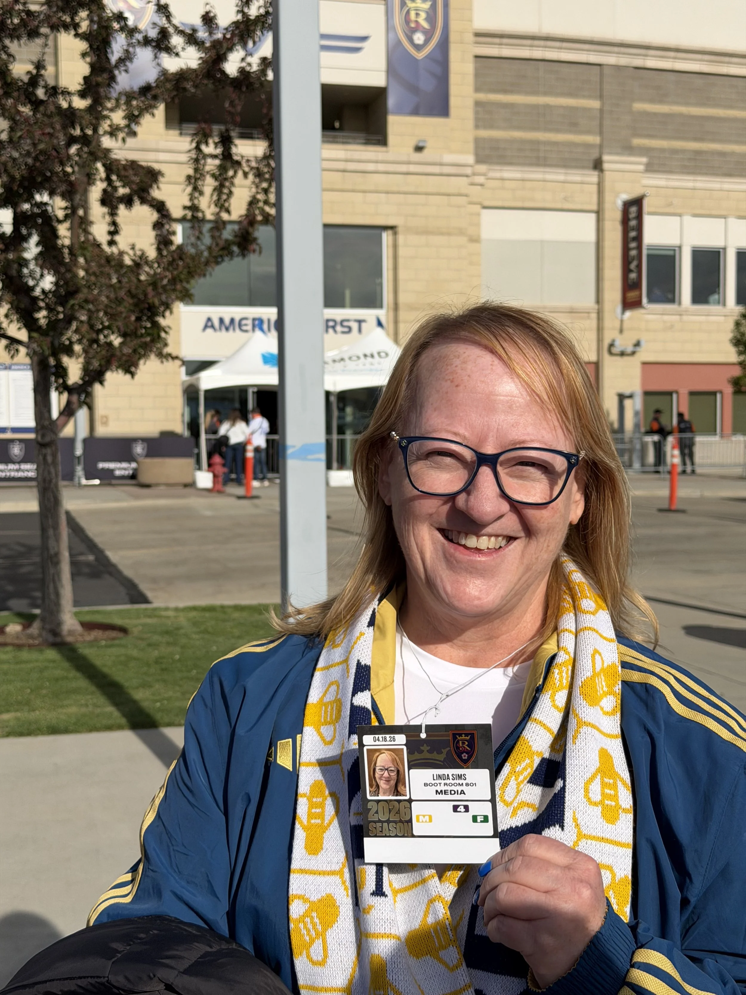 Linda Sims holding a media credential badge outside a stadium. She is wearing a blue jacket and a yellow and white scarf with the year 2026 season logo. The stadium background shows a signage reading 'American First' and some trees and people.