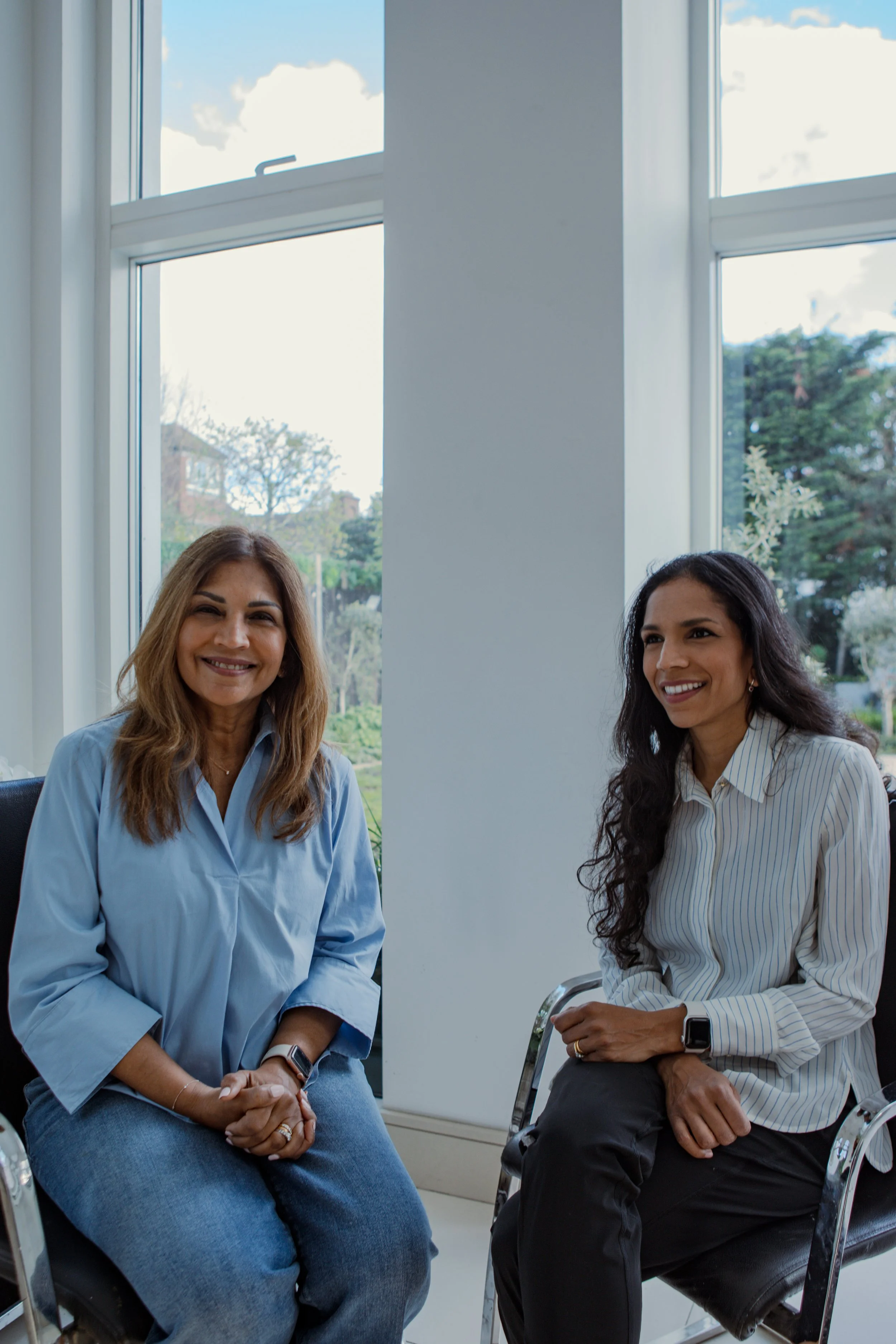 Two women sitting in chairs near a large window, smiling and engaging with each other.