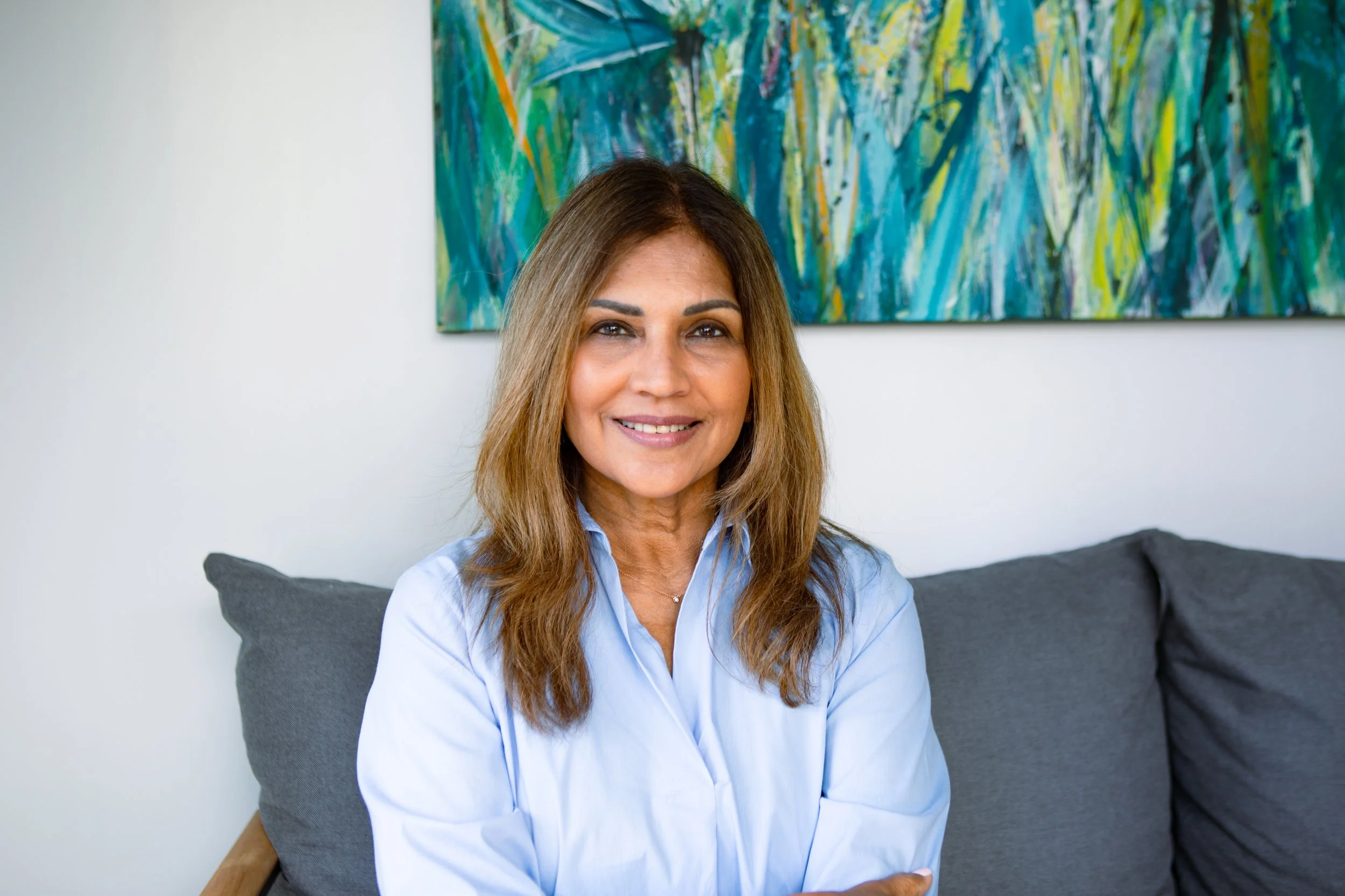 Smiling woman with long brown hair sitting on a gray couch in front of a colorful abstract painting.