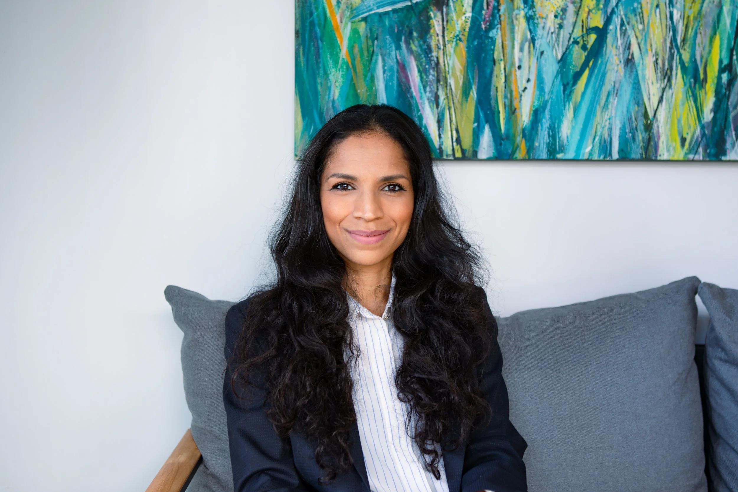 A woman with long dark hair smiling while sitting on a gray couch in front of a colorful abstract painting.