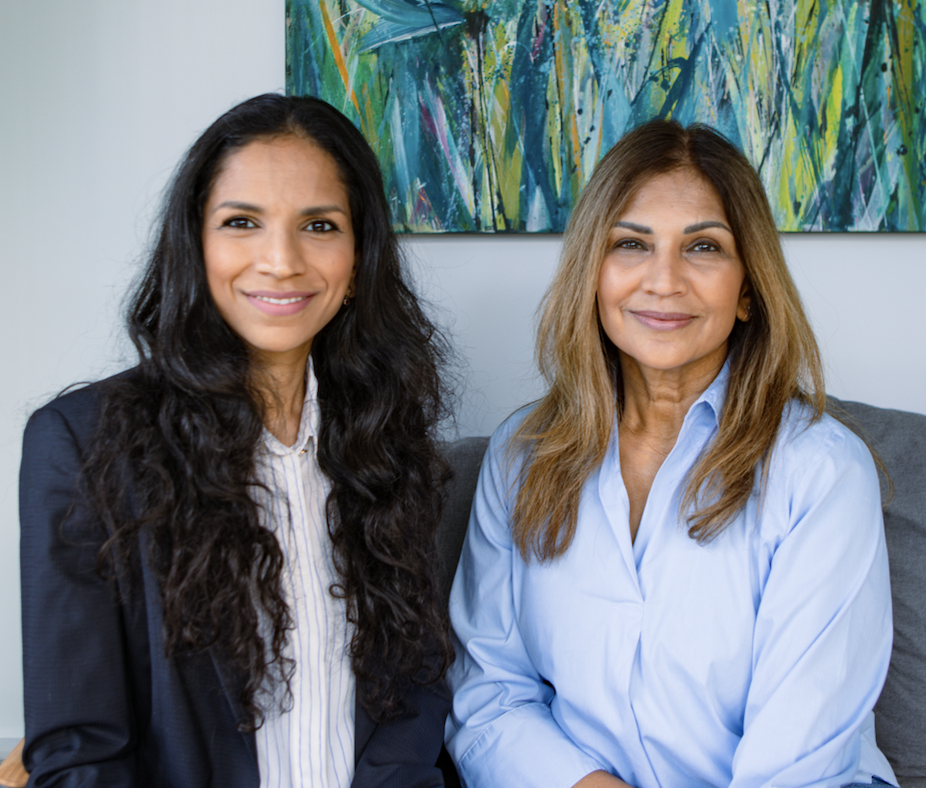 Two women sitting on a gray couch in front of an abstract colorful painting. The woman on the left has long, dark wavy hair and is wearing a dark blazer and white shirt. The woman on the right has shoulder-length light brown hair and is wearing a light blue button-down shirt.