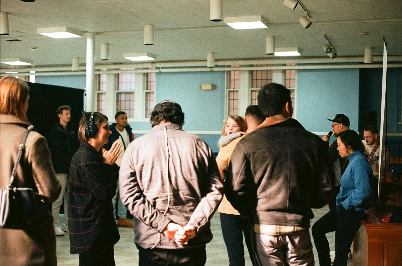 A group of people gathered indoors, some talking, some listening, with a woman holding a child, and a man speaking into a microphone.