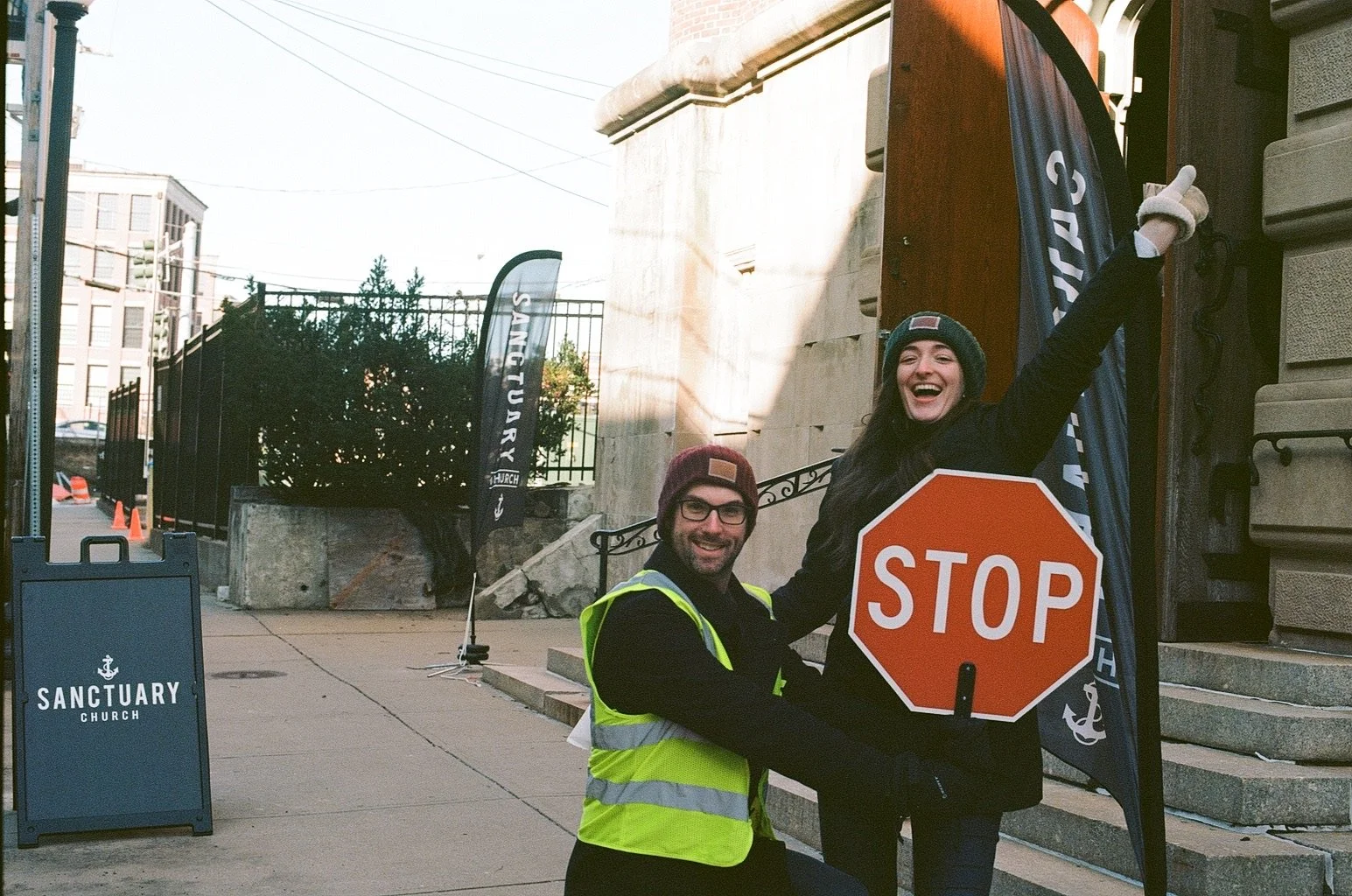 A smiling woman holding a stop sign, standing with a man wearing a reflective vest. They are on city steps near banners with the words 'Sanctuary Church' and a building with steps and an iron railing in the background.