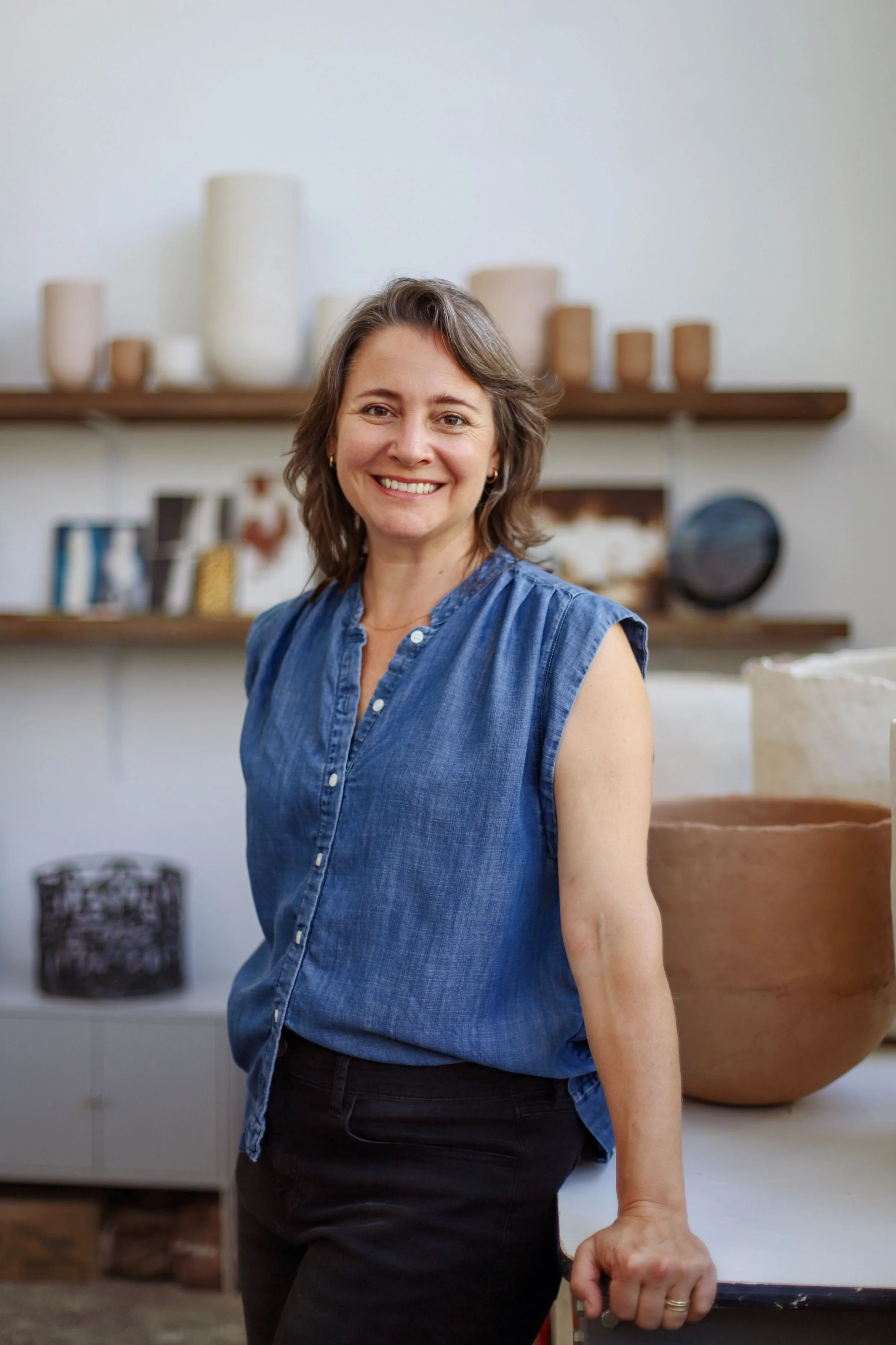 A woman with shoulder-length light brown hair, wearing a sleeveless denim shirt, smiling while standing indoors with shelves of pottery and books in the background.