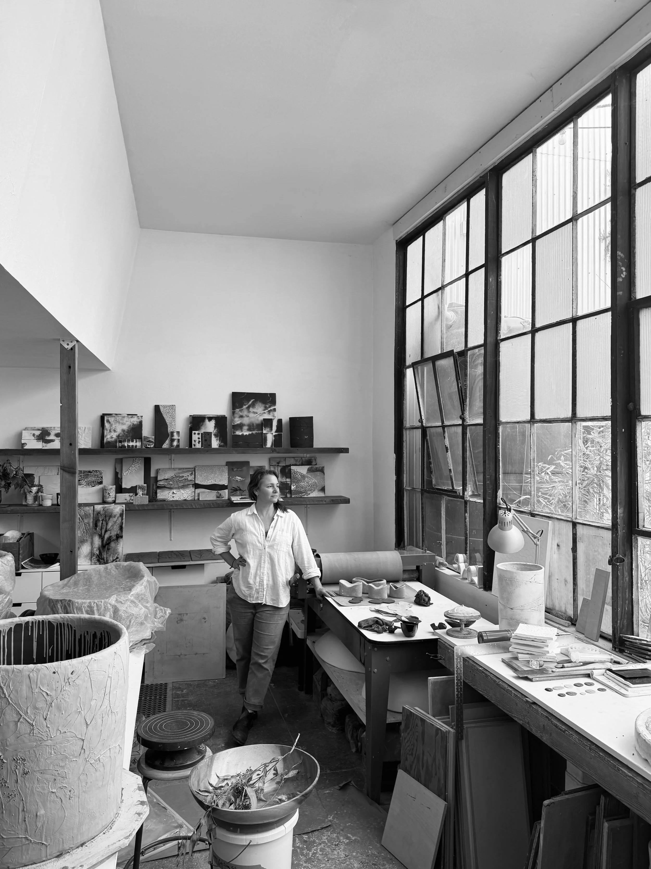 A woman standing inside an art studio surrounded by pottery and art supplies, with large windows on the right and art pieces on shelves behind her.