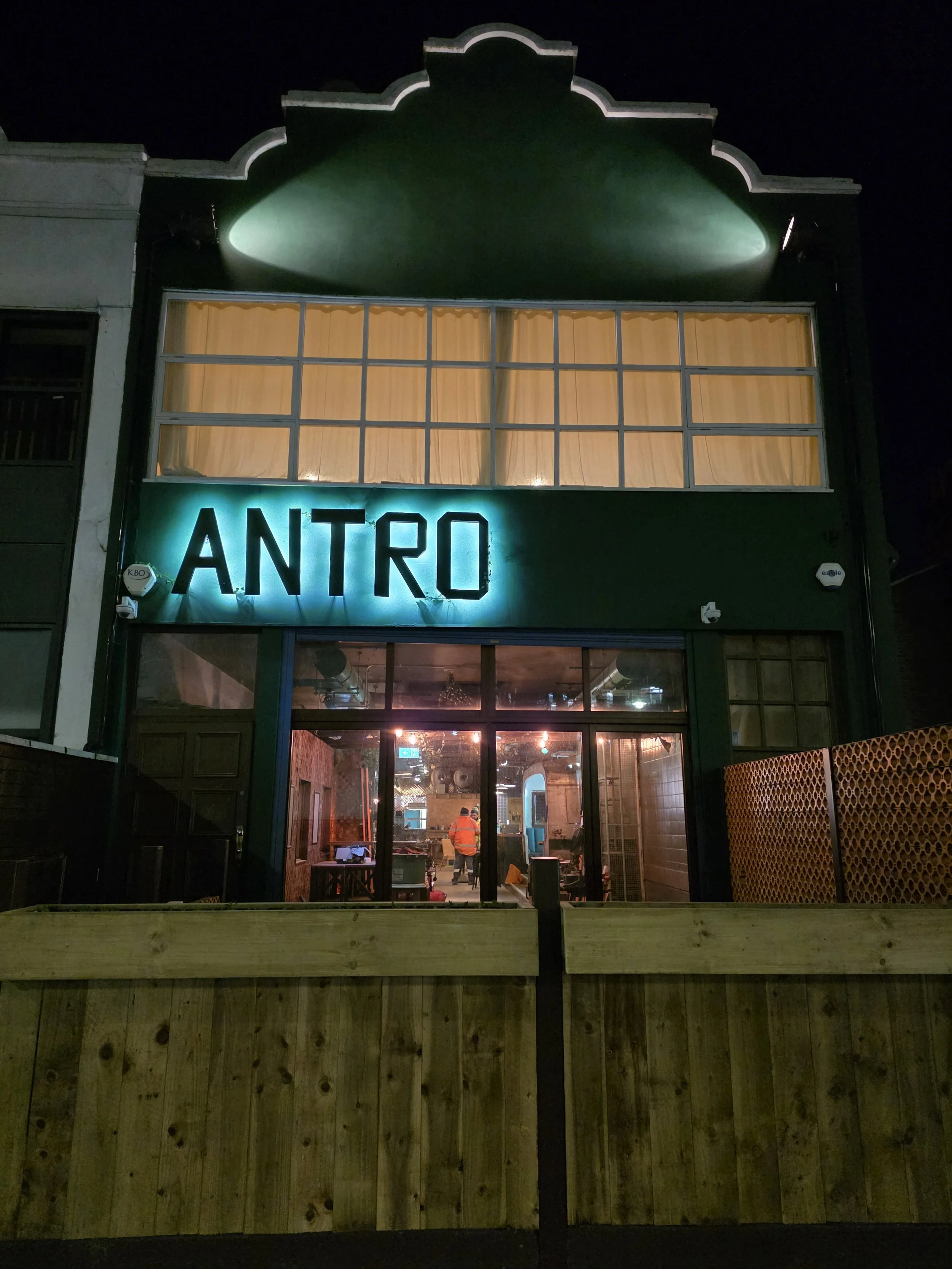 The exterior of a building at night with a green facade and a large illuminated sign that reads 'ANTRO'. The building has large windows with curtains, and the interior is visible through the glass, showing warm lighting and people inside.