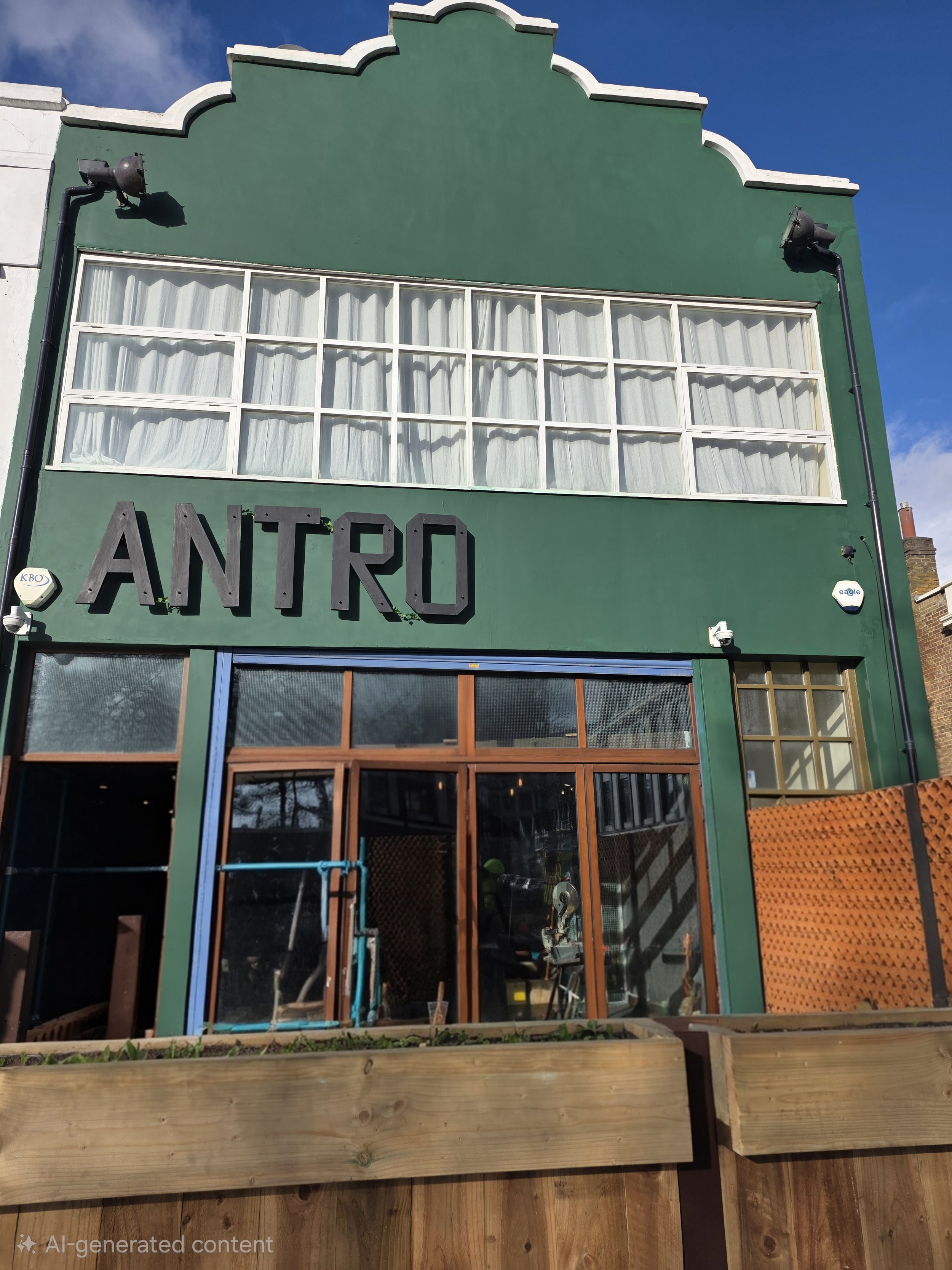 Front of a green building with large windows, black sign spelling 'ANTRO', and a wooden patio area in front.