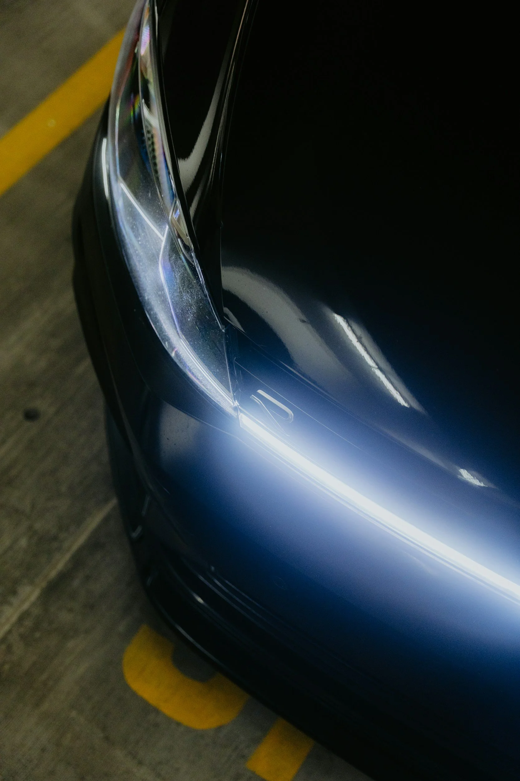 Close-up van de koplamp en motorkap van een zwarte auto in een parkeerplaats.