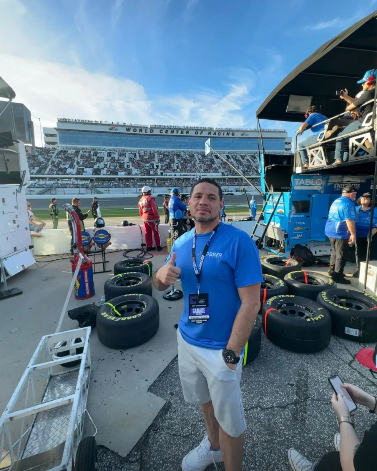 Man standing in a racing garage area with racing tires behind him, race track and stands in the background, giving a thumbs-up.