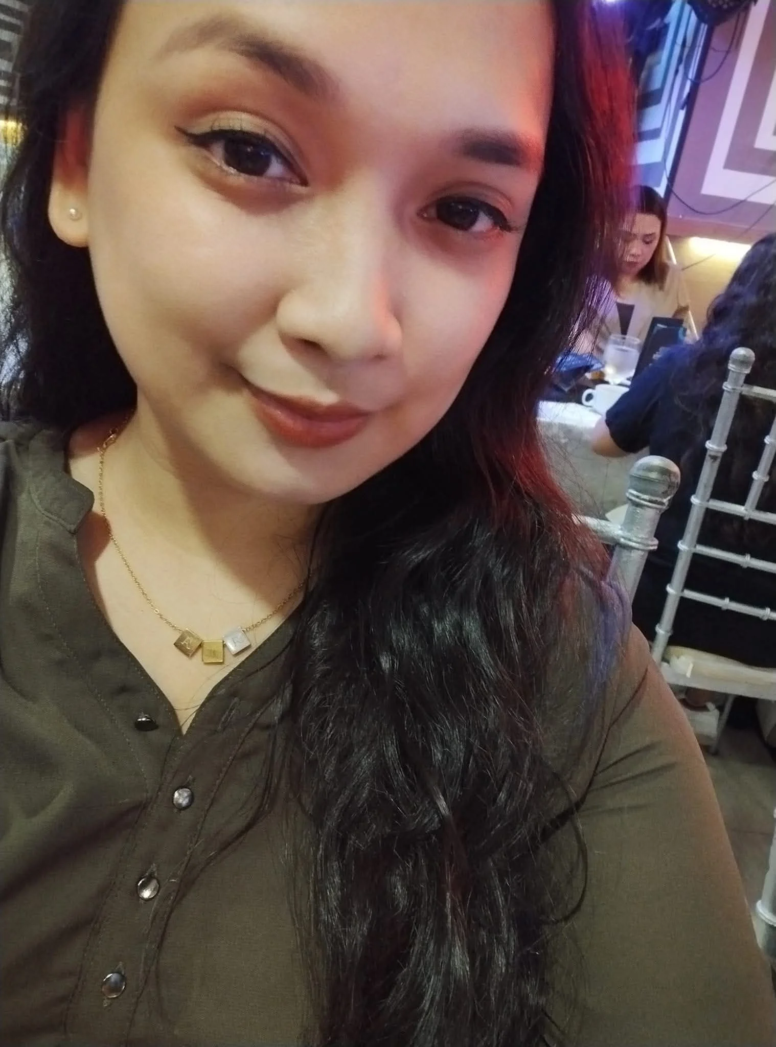 Close-up of a woman with long dark hair, wearing a brown shirt and a gold necklace, smiling at the camera in a restaurant setting with other patrons in the background.