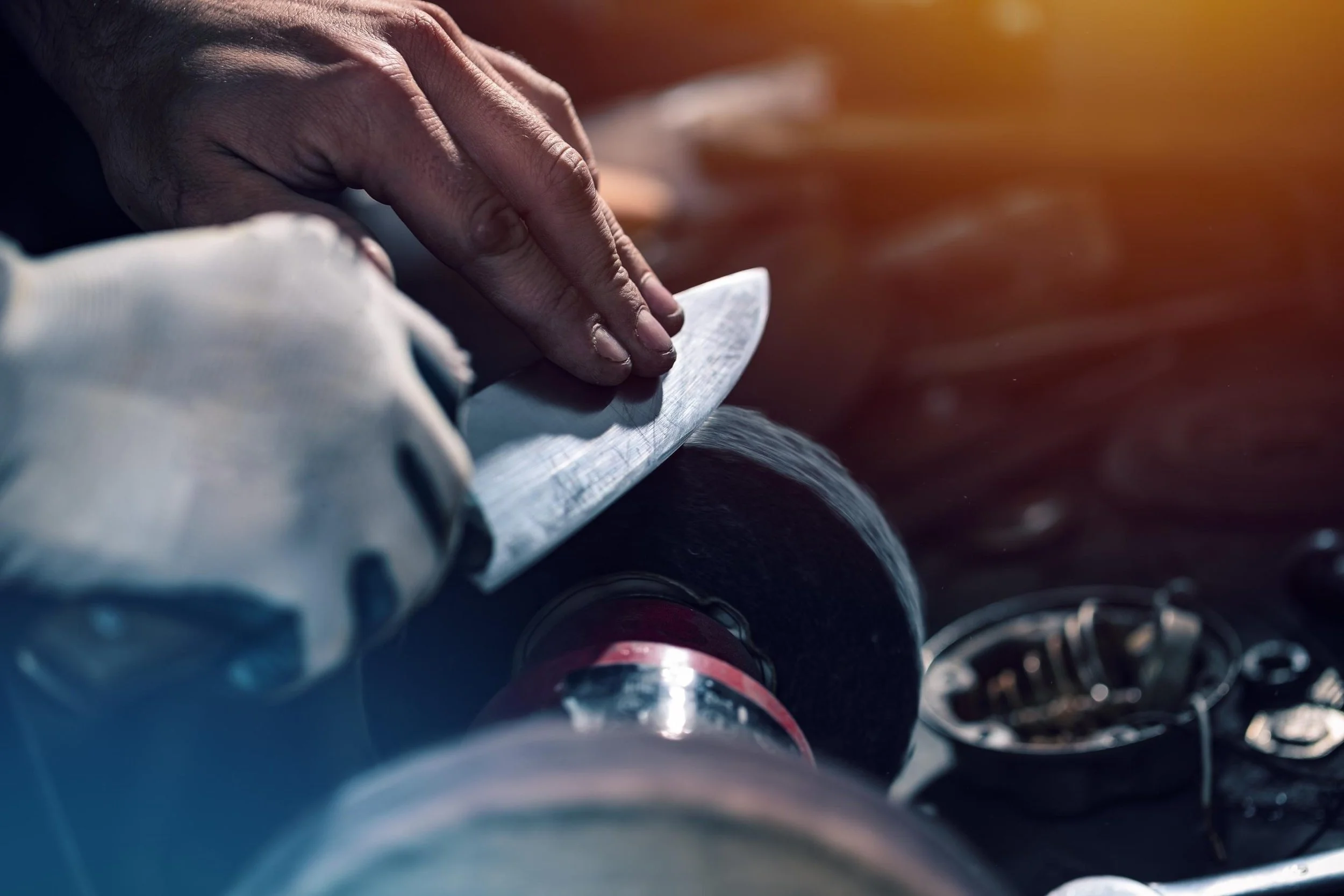 A person sharpening a knife with a portable electric grinder. The person's hands are visible, and they are wearing a light-colored long-sleeved shirt. The grinder is black with a red band, and there are some metal objects and the grinder's cord nearby.