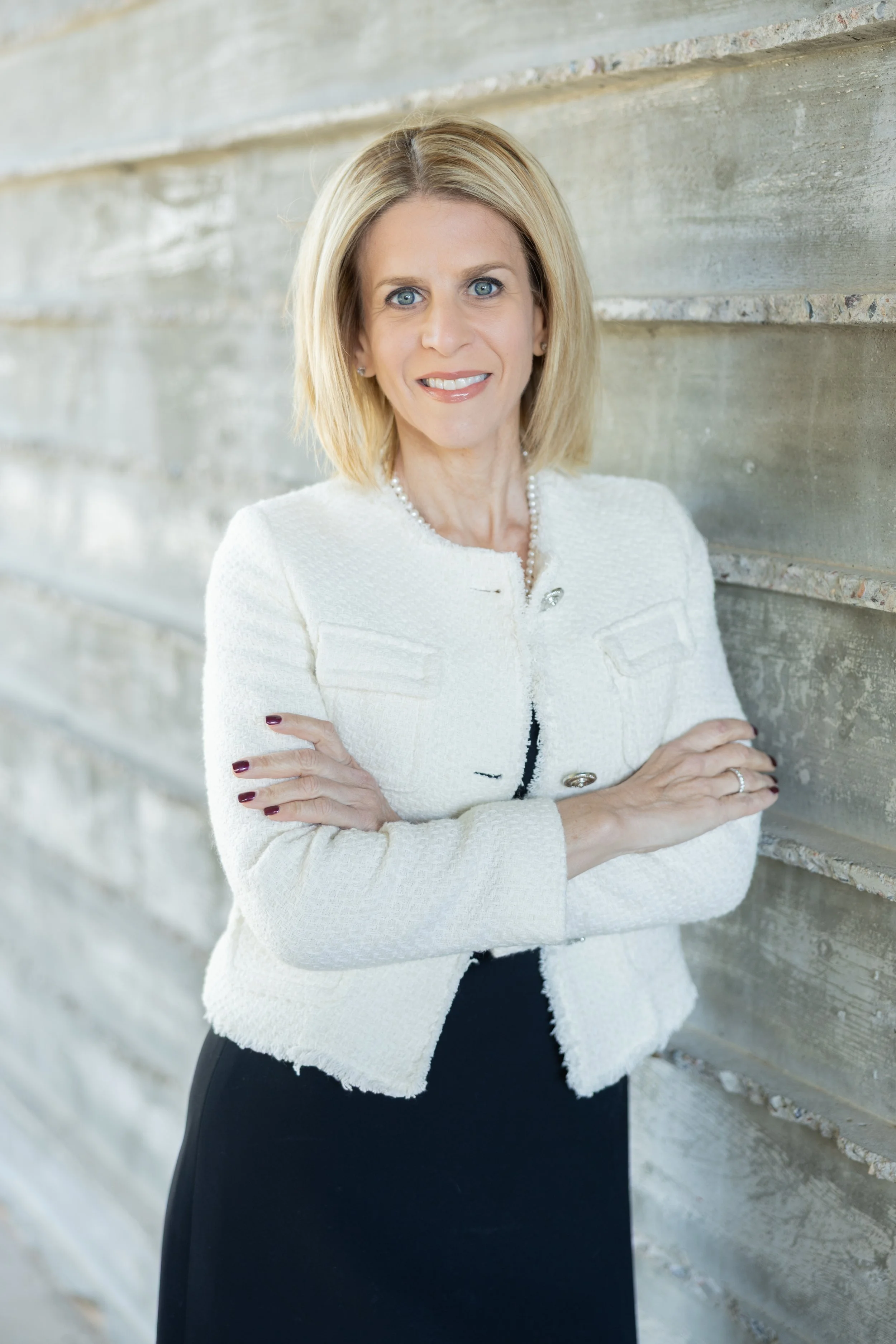 A woman with blonde hair and blue eyes standing against a rustic wooden wall, wearing a white jacket and black dress, with her arms crossed, smiling.