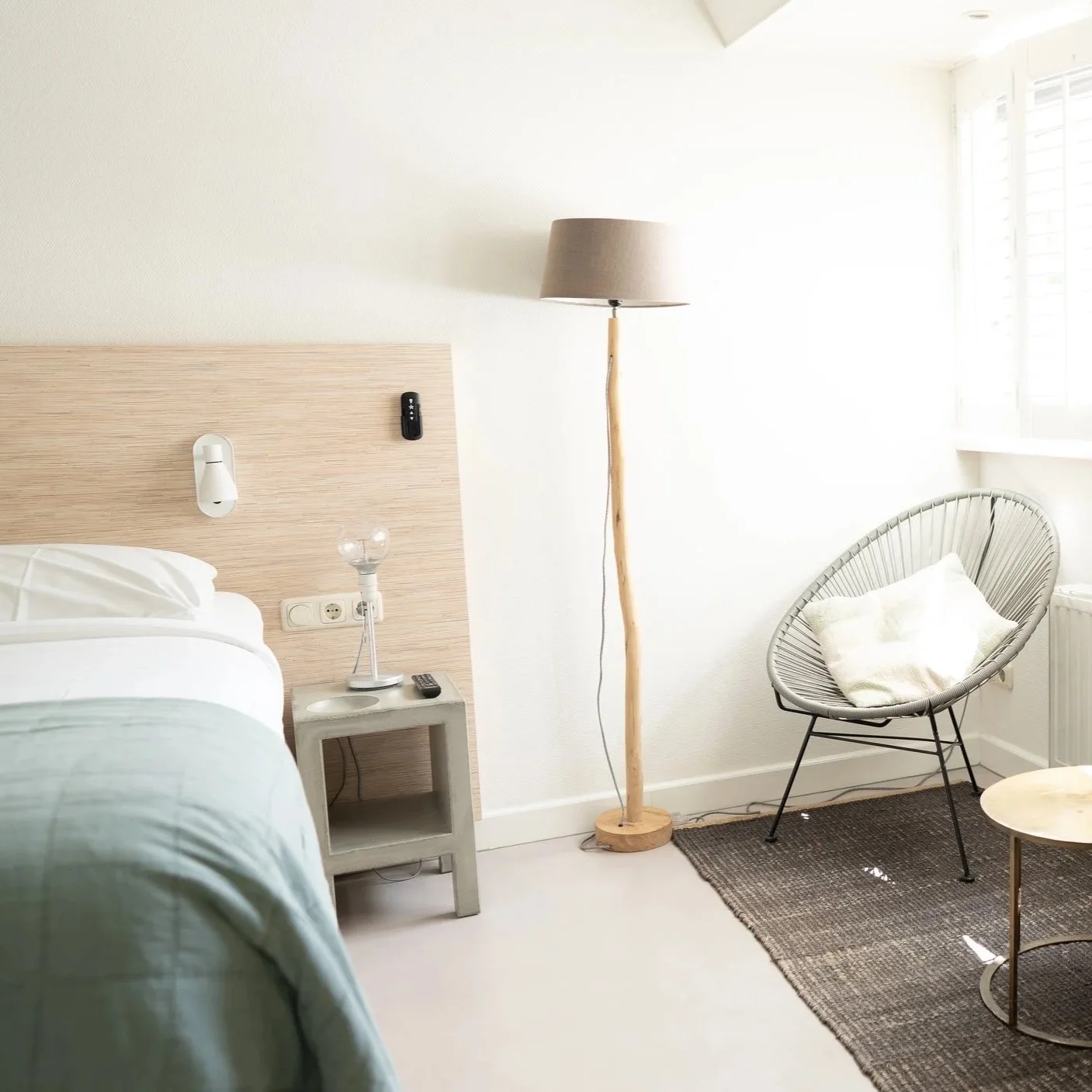 Kamer met bed, nachtkastje, lamp, relaxstoel met kussen, vloerkleed en groot raam met jaloezieën.