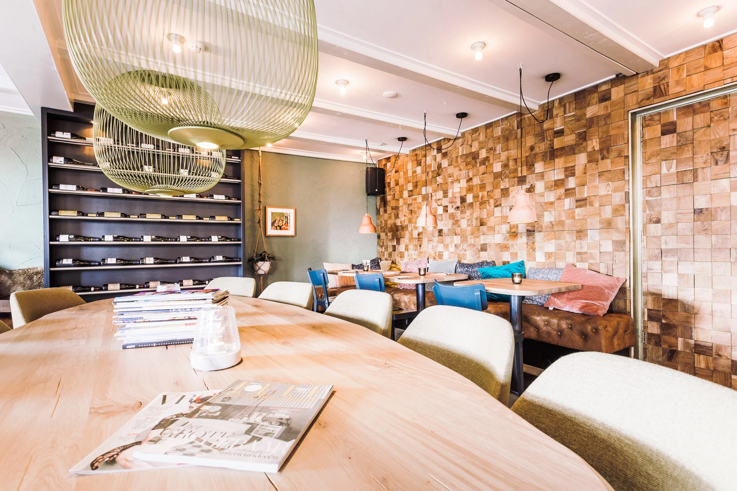 Interieur van een restaurant met een groot houten tafel, stoelen, een muur met een houten panelen en een bank met kussens. Hangers en spiegels hangen aan het plafond, en er is een plank met wijnflessen.