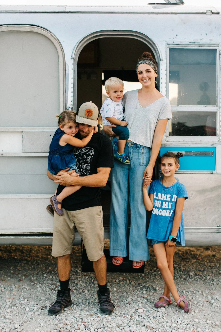 A family of five standing in front of a vintage camper trailer, smiling and holding hands, with one adult holding a child and another child leaning on her. The scene appears to be outdoors on a gravel surface.