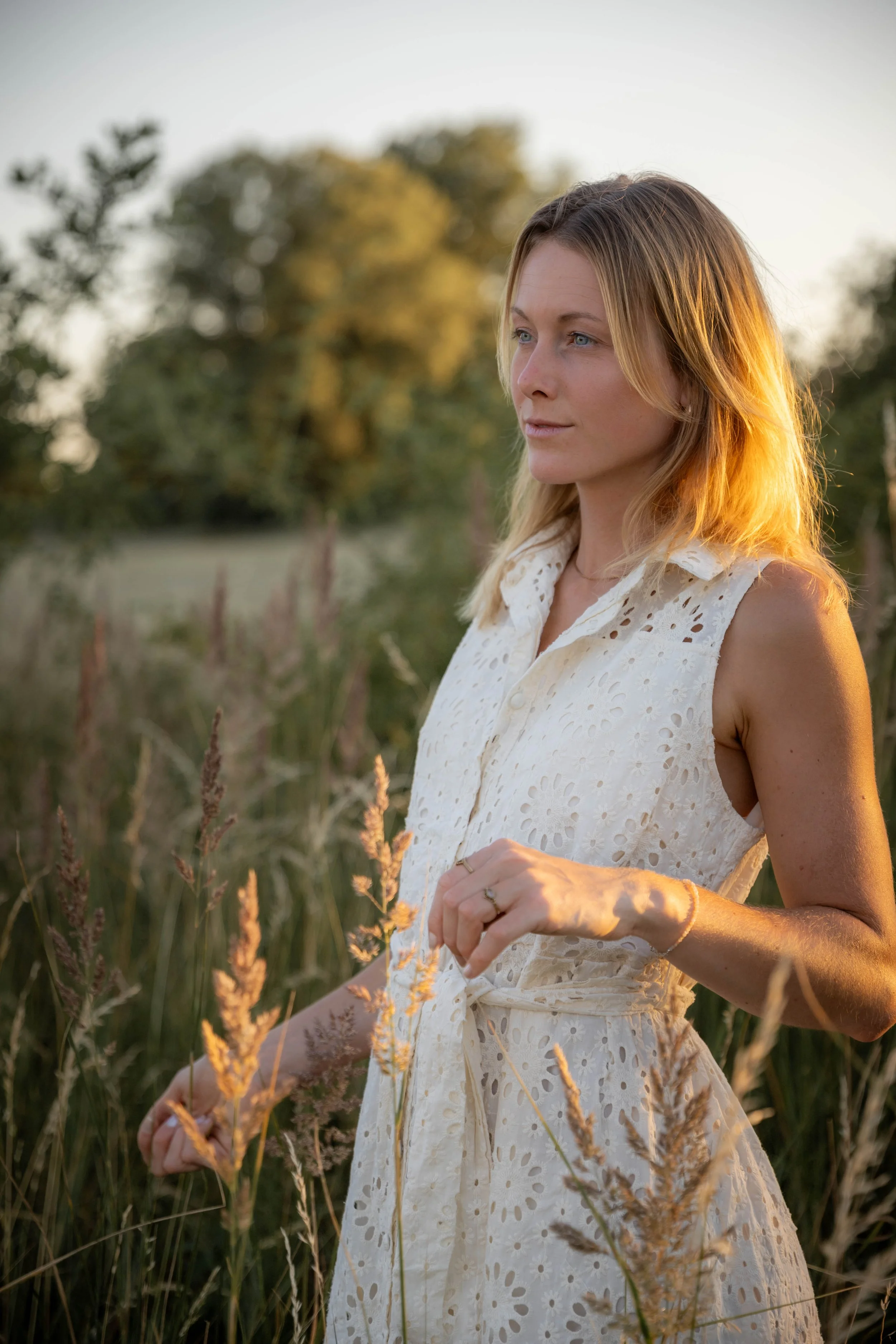 A woman stands in a field of tall grasses during sunset, wearing a white, sleeveless, eyelet dress, with blonde hair and a gentle expression.