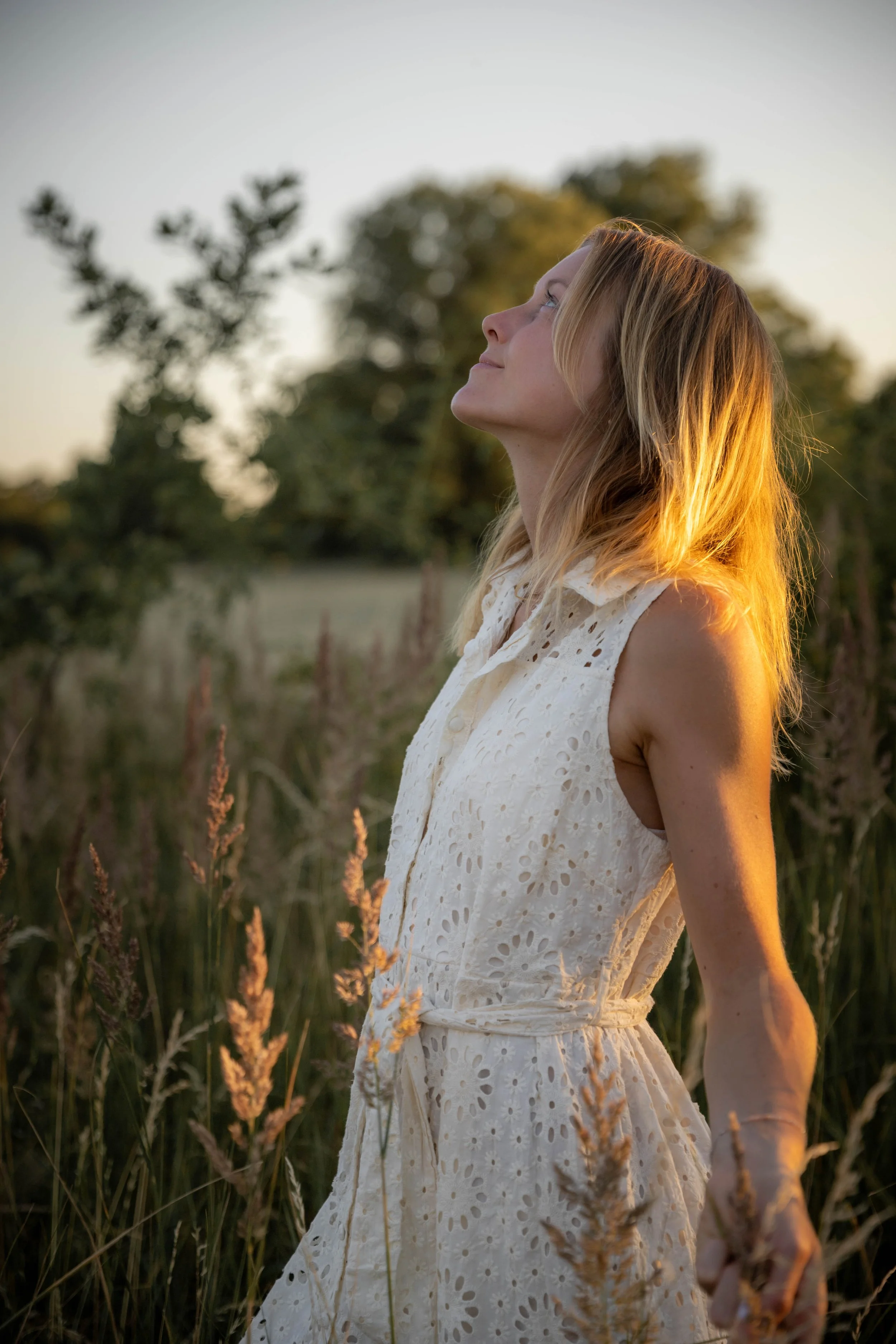 A woman with blonde hair wearing a white sleeveless dress standing in a field of tall grass and plants during sunset, with trees in the background and her face turned upwards.