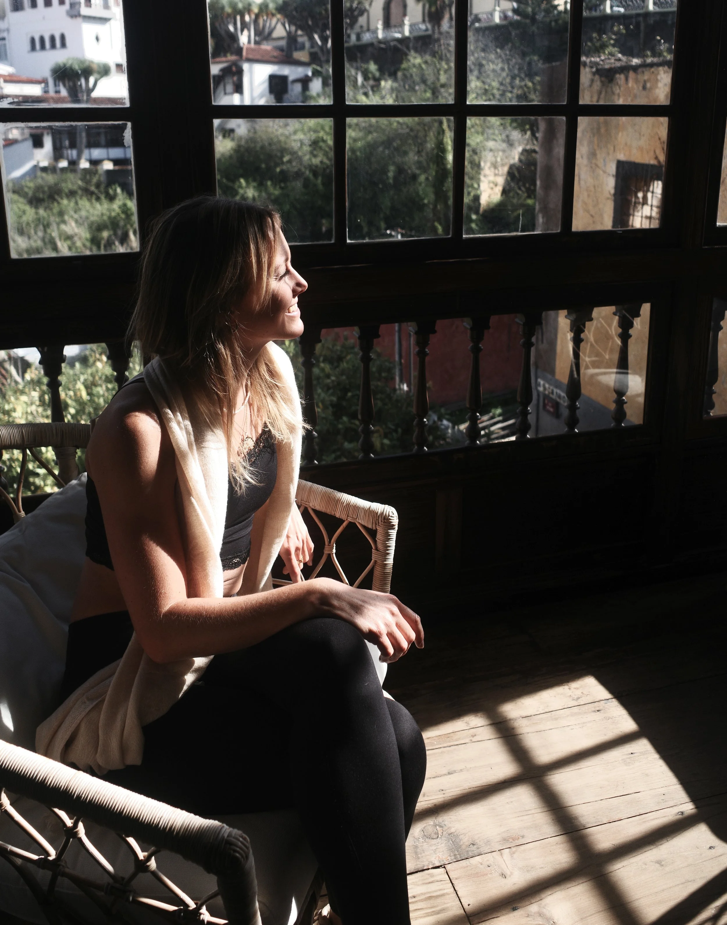 A woman sitting on a wicker chair near large windows, enjoying sunlight and a view of trees and buildings outside.
