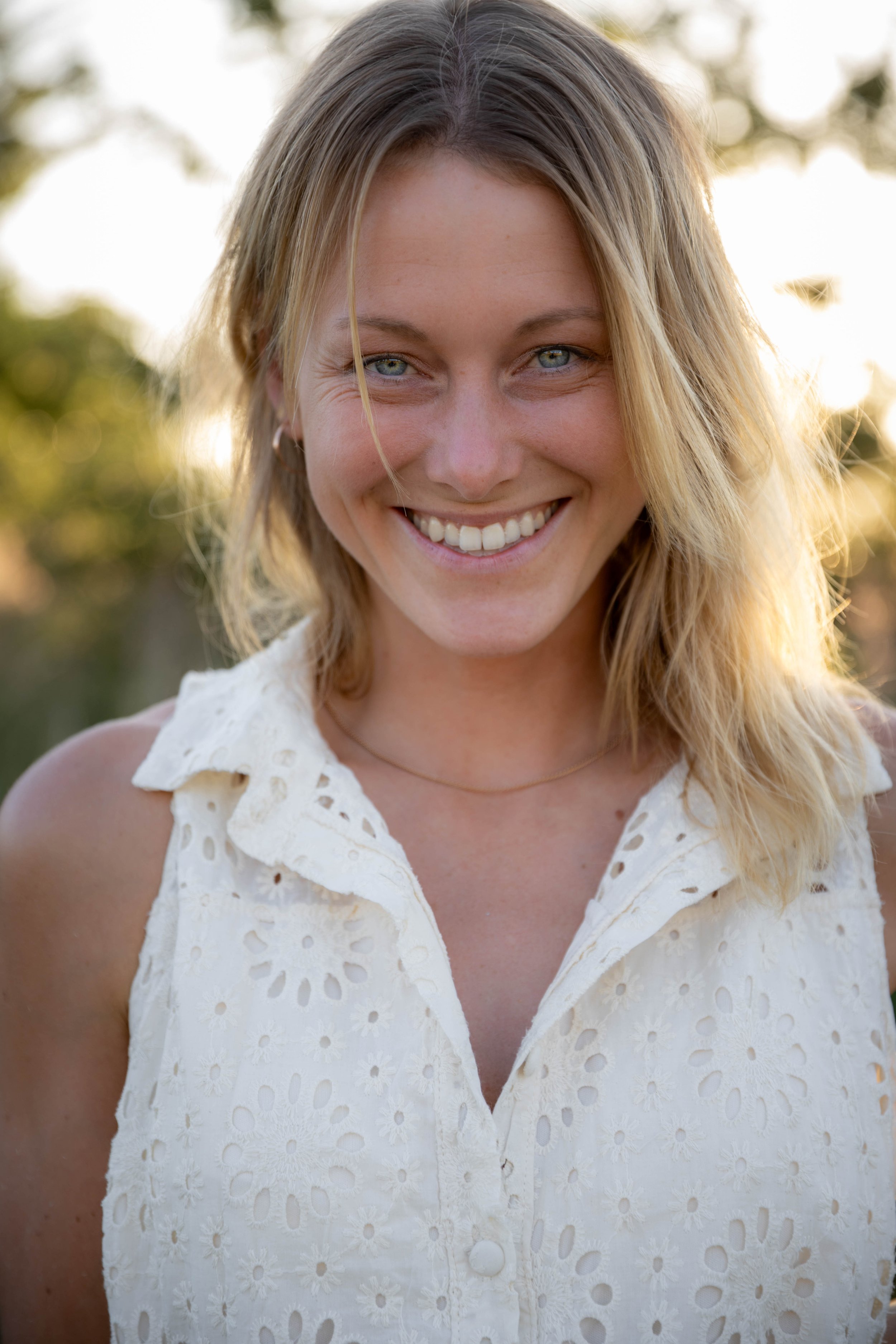 Close-up of a young woman with blonde wavy hair smiling outdoors at sunset, wearing a white eyelet sleeveless shirt.