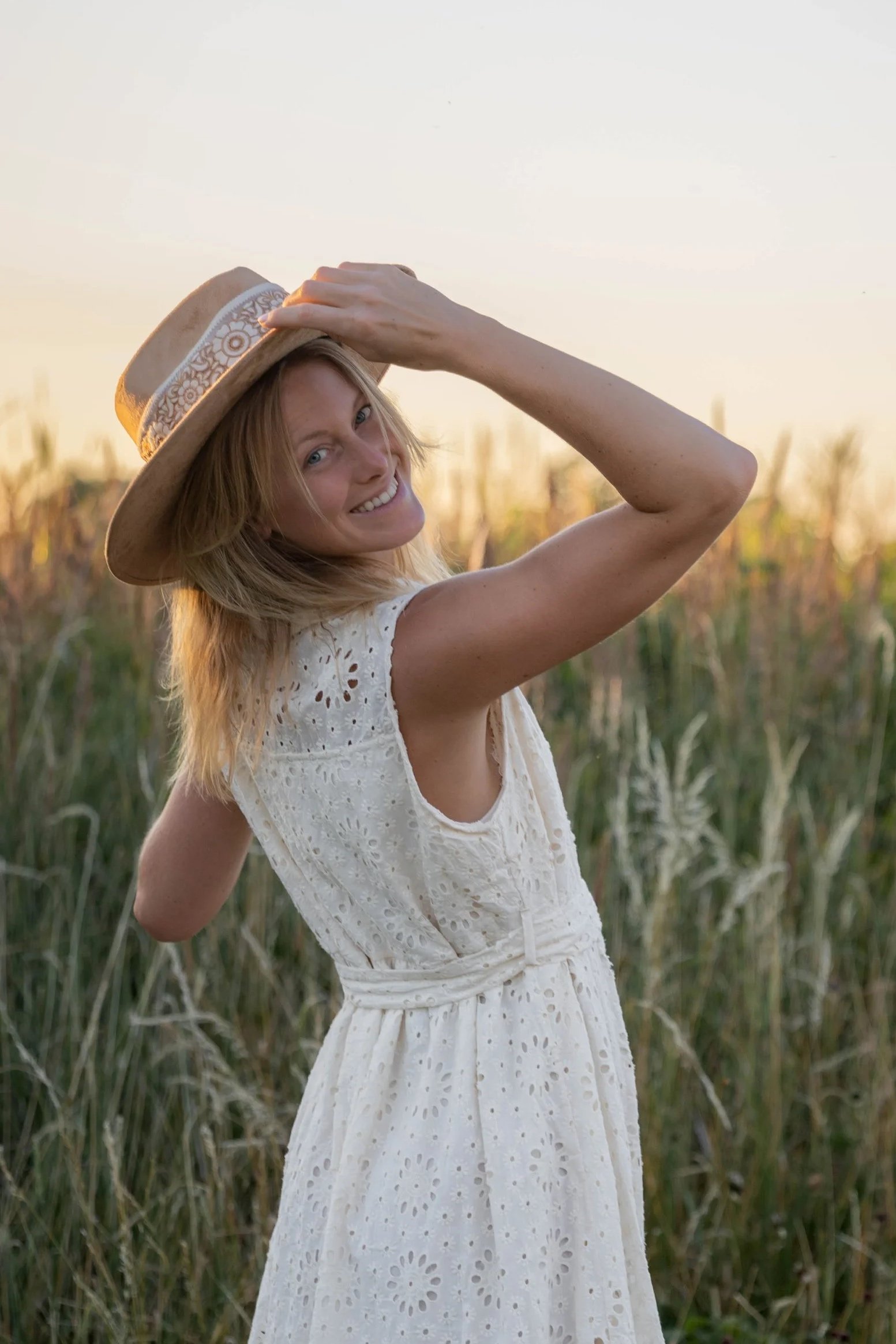 A woman smiling outdoors in a white eyelet dress and a wide-brimmed straw hat, standing in a field of tall grass with a soft sunset in the background.
