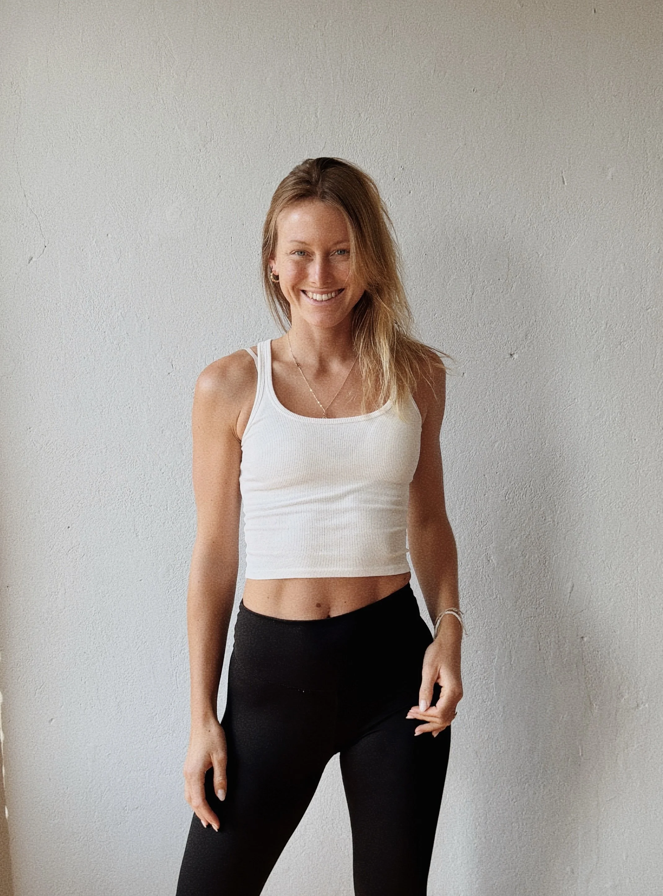 A woman with long red hair, smiling, wearing a white sleeveless crop top and black high-waisted pants, standing against a plain white wall.