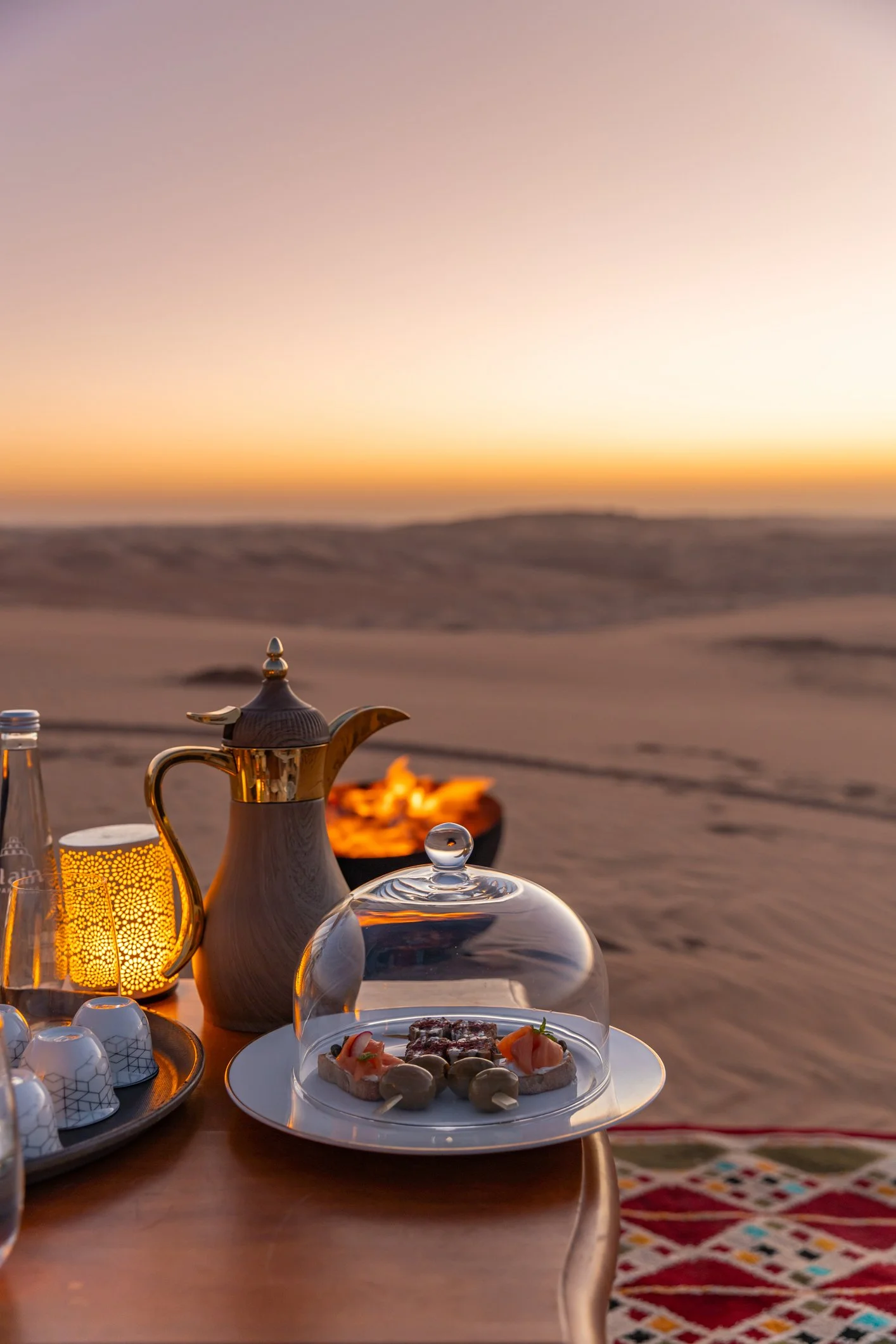 An elegant outdoor dining setup on a desert landscape at sunset, featuring a table with a glass dome covering a plate of appetizers, a teapot, a candle holder, and a tray of small cups, with a blurred background of sand dunes.