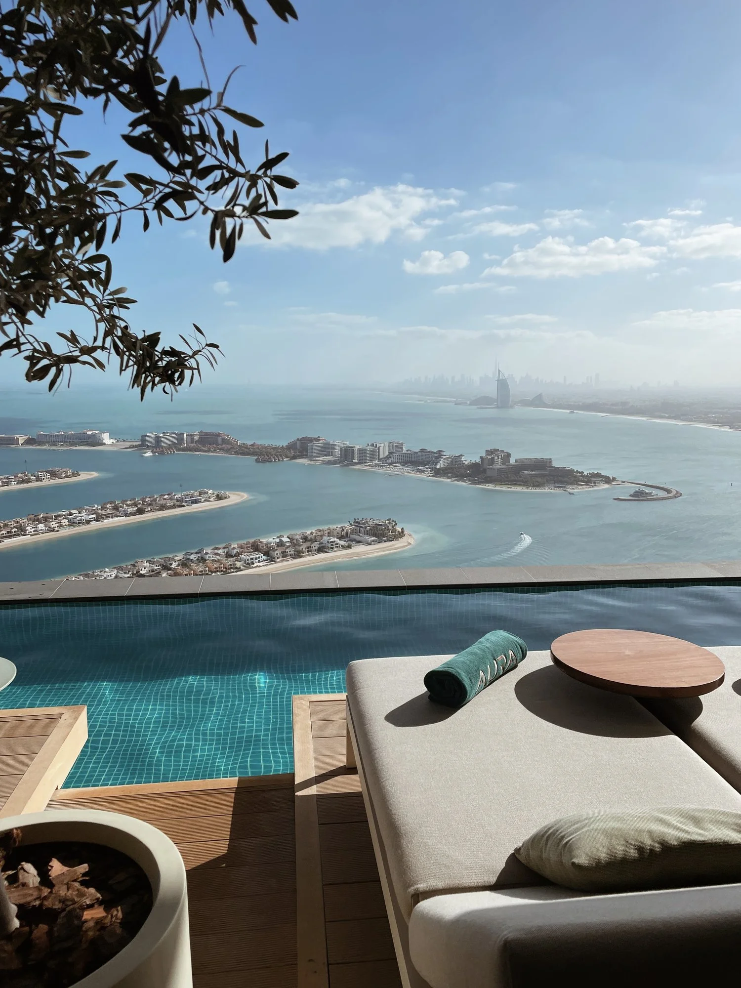 View of an infinity pool overlooking Dubai skyline with the Burj Al Arab in the background, seen from a terrace with lounge chair, rolled towel, and small round table.