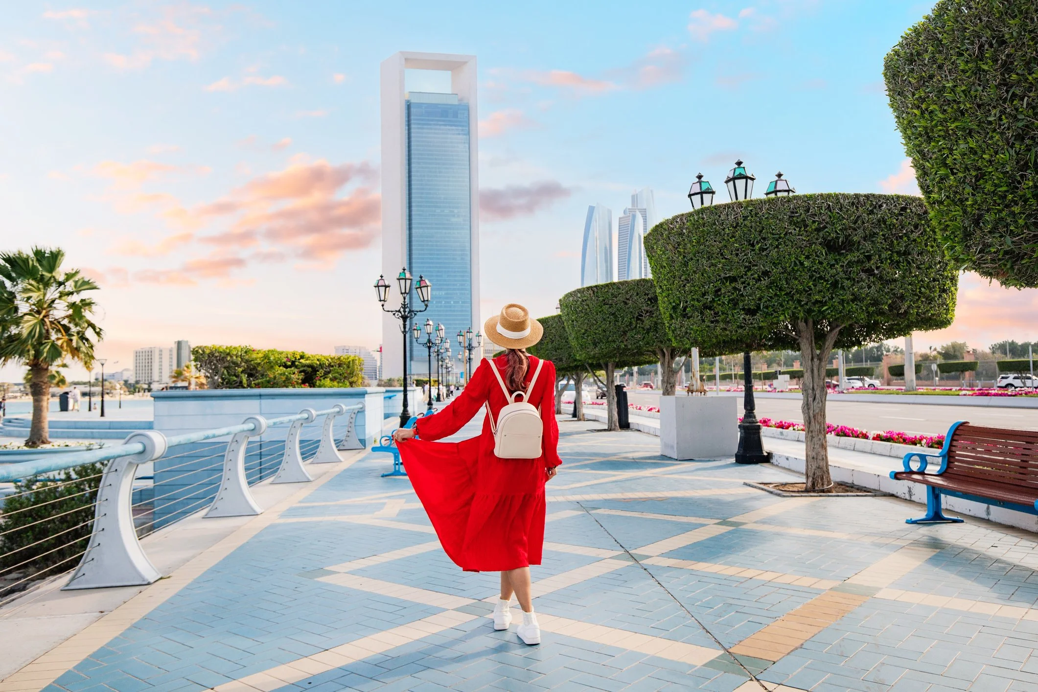 A woman in a red dress and straw hat walking along a coastal promenade with modern high-rise buildings and a partly cloudy sky in the background.
