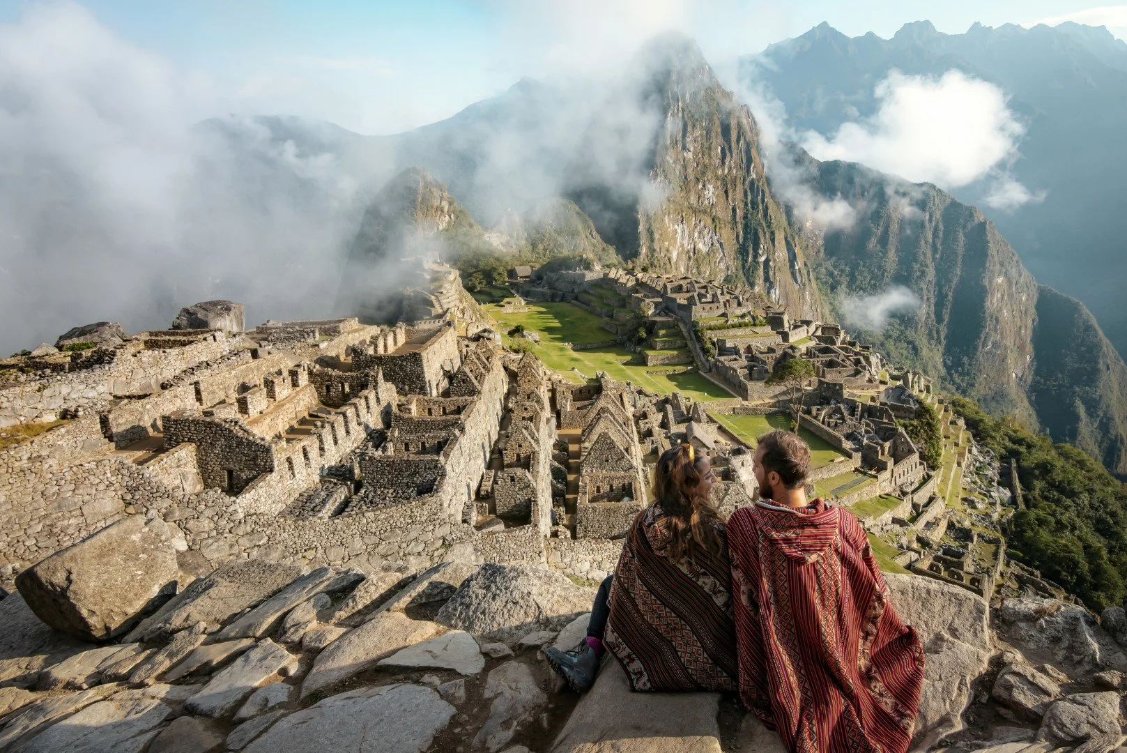 A couple sitting on rocks overlooking Machu Picchu, an ancient Incan citadel in the Andes mountains, with clouds and steep mountain peaks in the background.