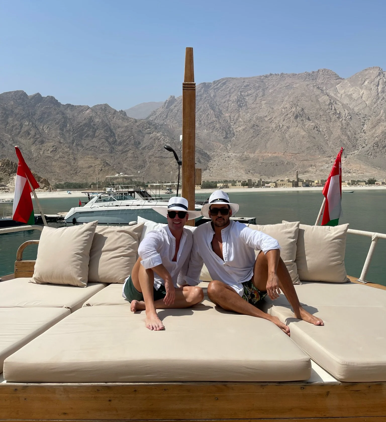 Two men sitting on a cushioned lounge on a boat with two red and white flags, water, boats, mountains, and a clear sky in the background.
