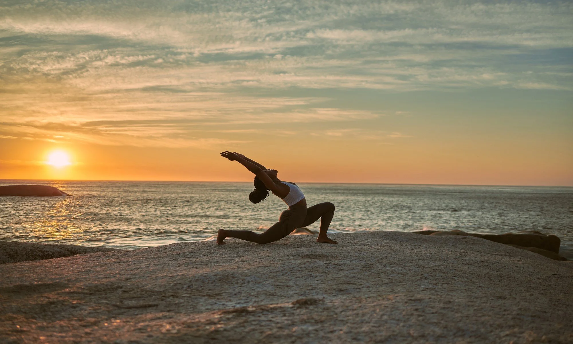 A woman practicing yoga on a large rock at the beach during sunset, stretching her arms overhead in the warrior pose.