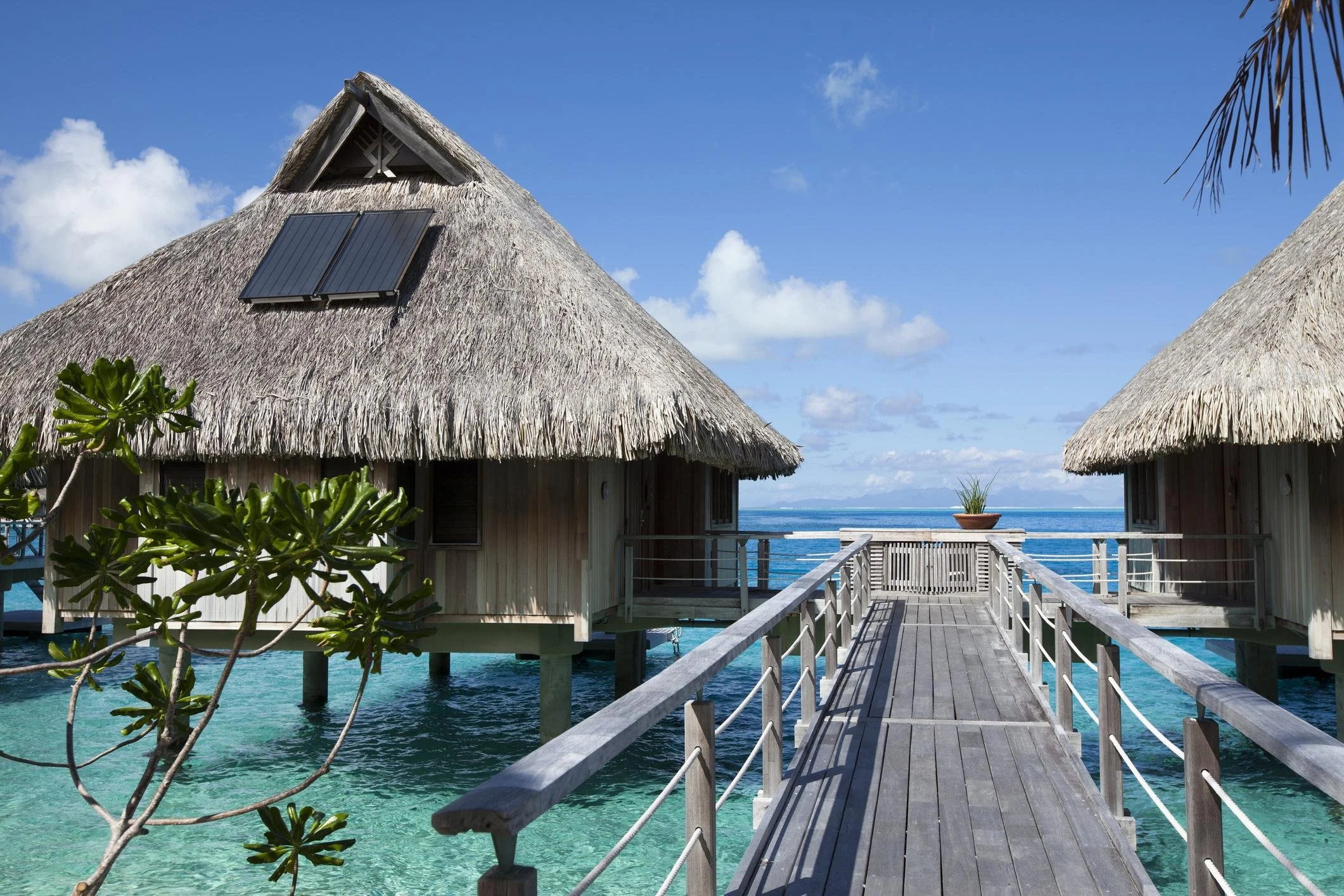 Overwater bungalows with thatched roofs and a wooden walkway on clear blue water, under a partly cloudy sky.