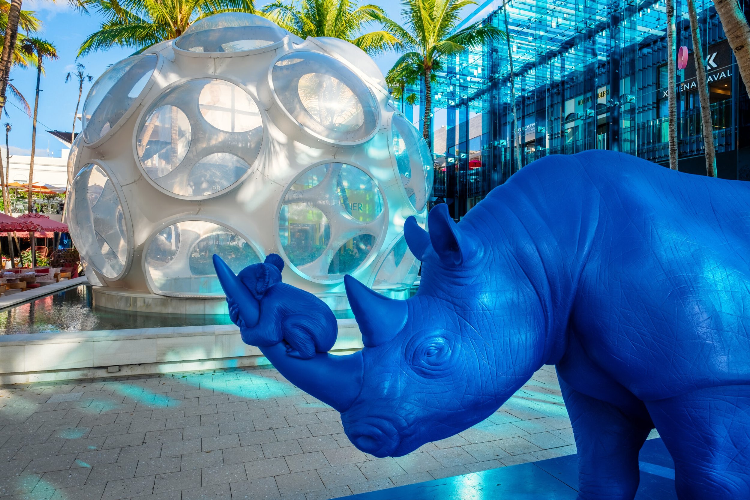 A blue elephant sculpture holding a small ball with its trunk in front of a futuristic glass spherical structure and palm trees in the background.